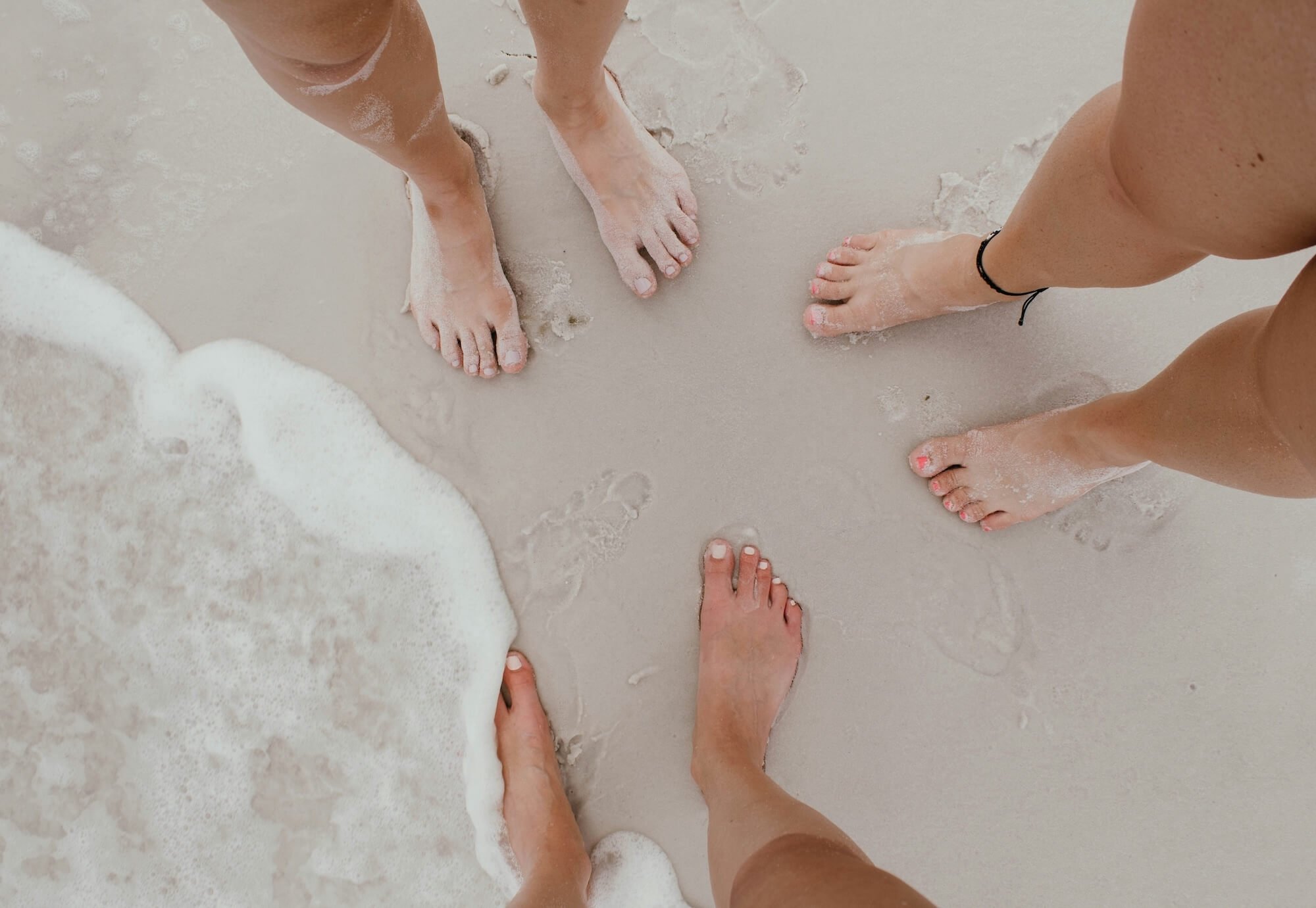 Group of women standing barefoot together in the sand at the water's edge, representing shared community and connection