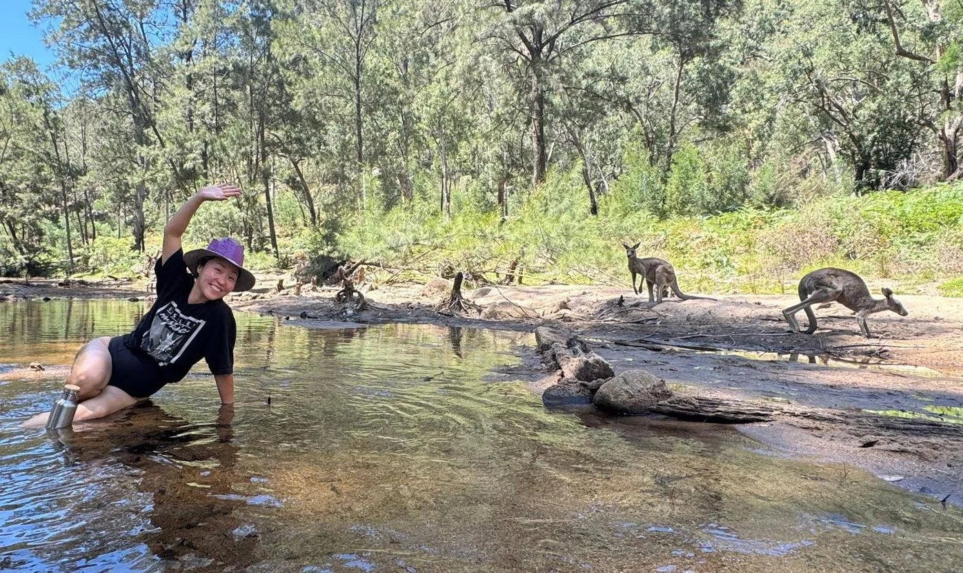 Woman in a black t-shirt and purple hat smiling and waving while crouching in a shallow river, with three kangaroos on the riverbank in a forested area.