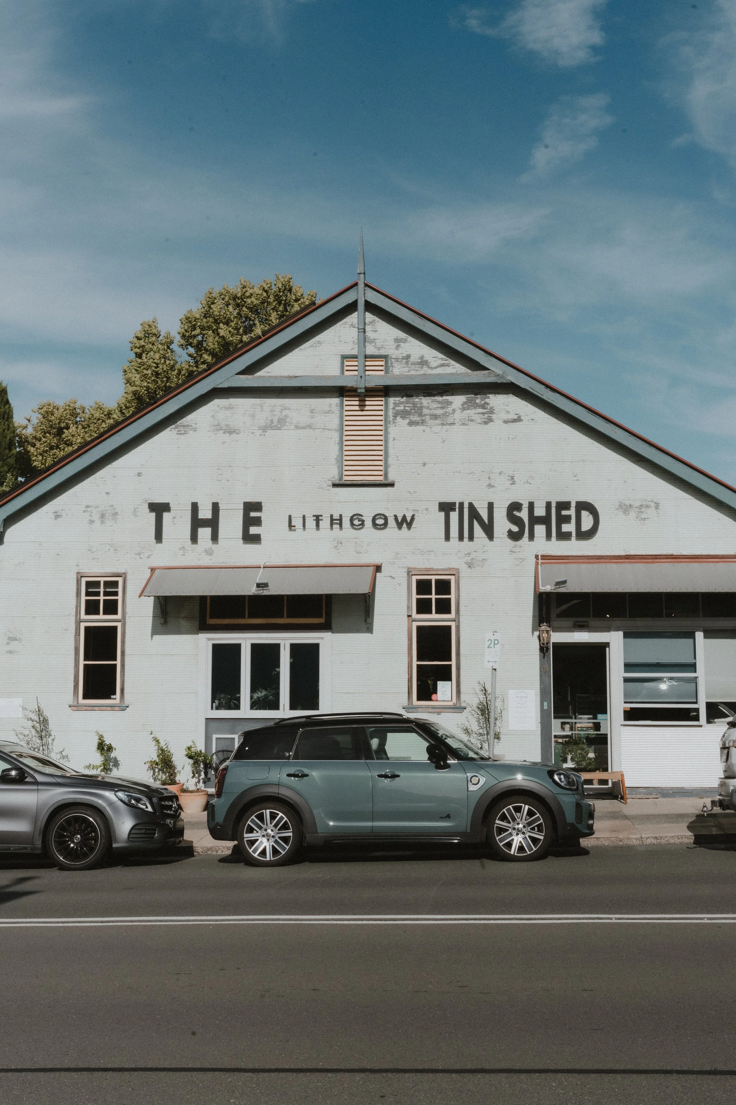 Front view of a white building with signs that read 'The Lithgow Tinshed' and a parking lot with two cars parked in front.