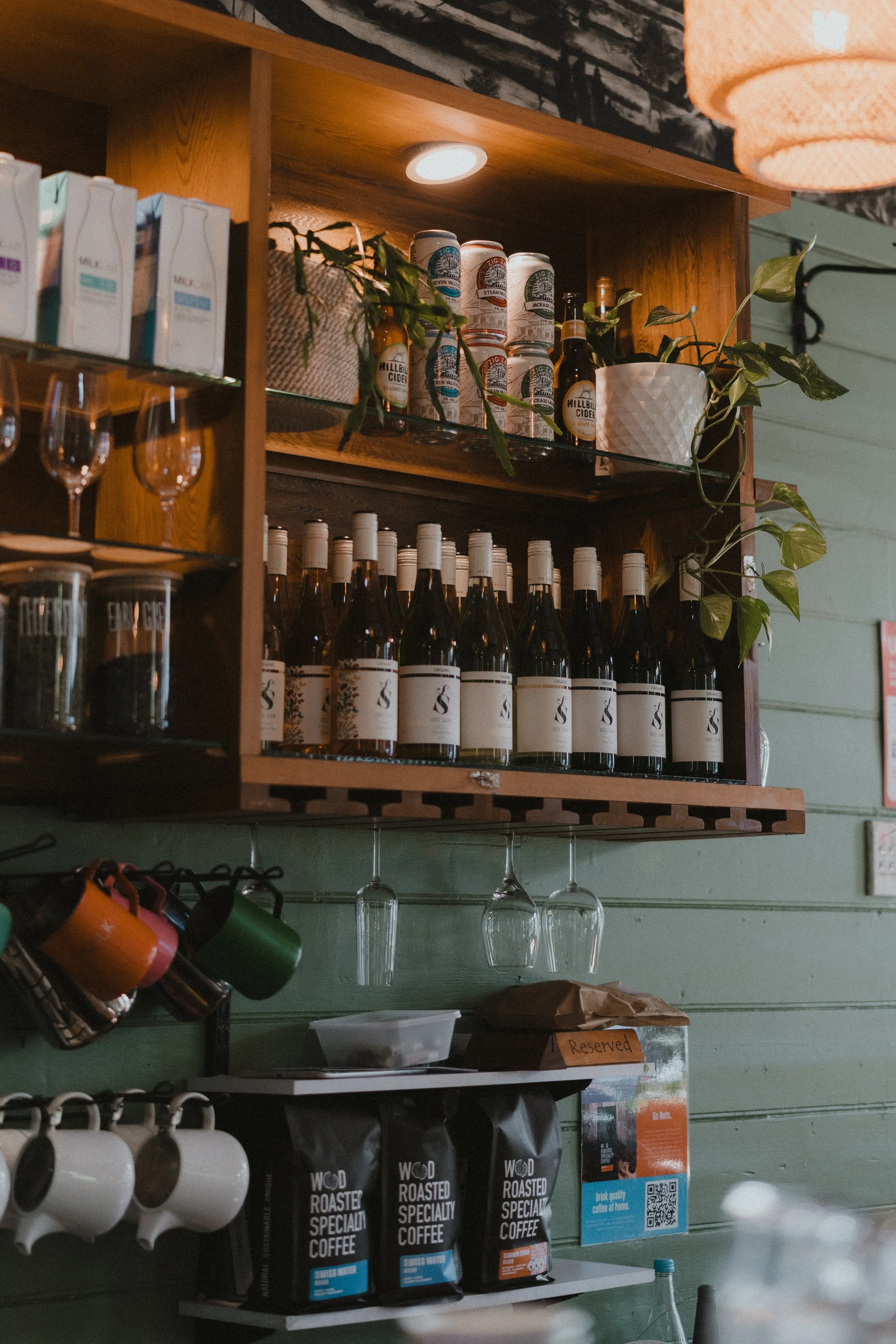 A kitchen or cafe shelf displaying various beverages, coffee mugs, glasses hanging upside down, coffee bags, and potted plants.