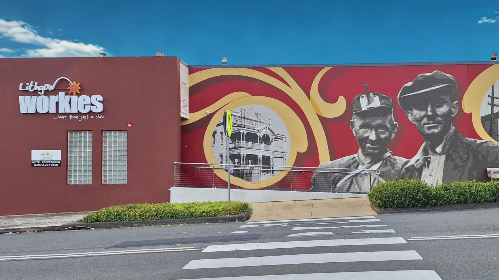 A mural on a building featuring two men in grayscale, one wearing a racing helmet. The building's logo reads 'Lithgow workies' with a tagline 'More than just a club.' A circular illustration of a historic building is also part of the mural. The scene includes street elements such as a crosswalk, bushes, and a small sidewalk.