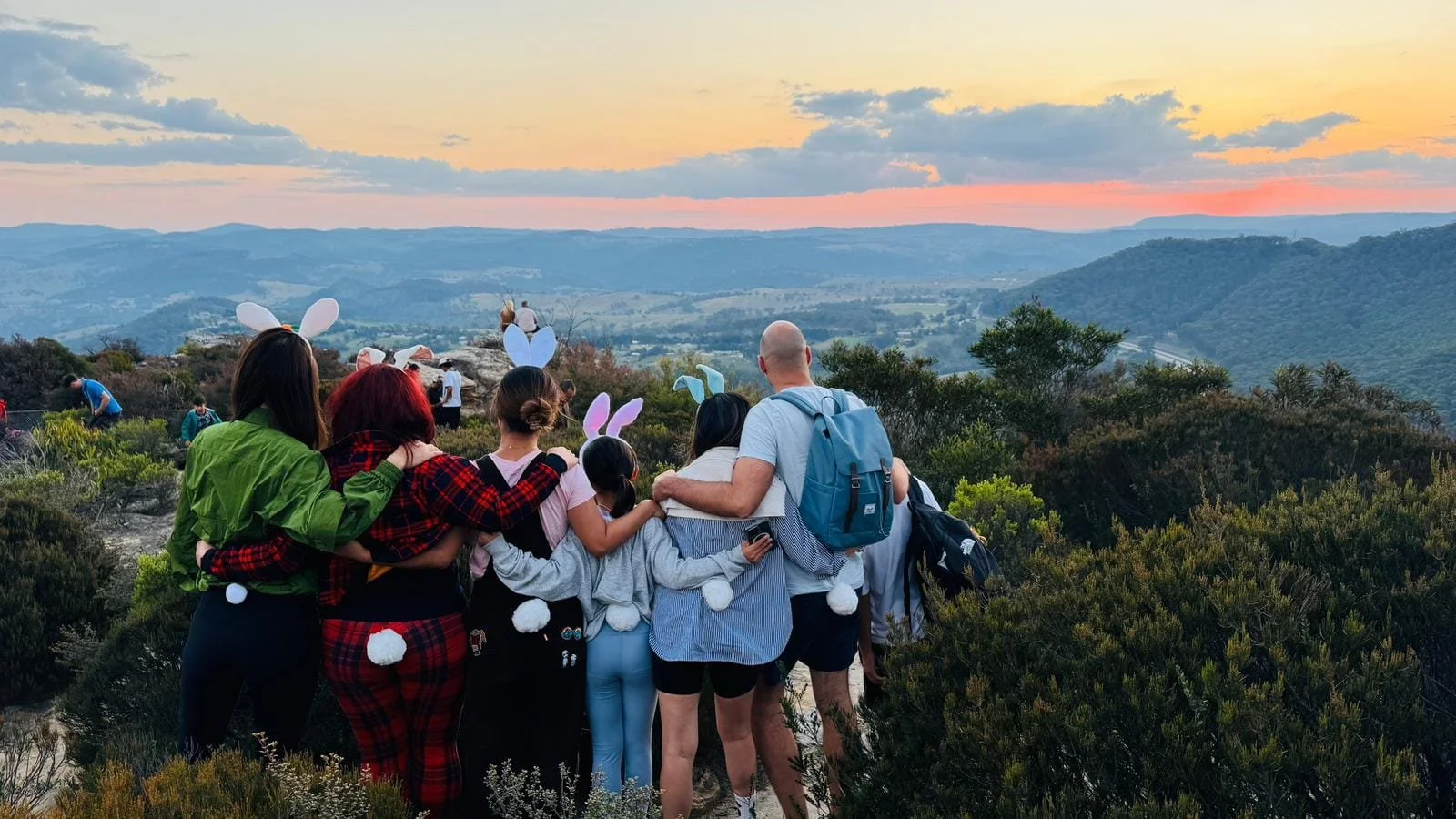 Group of people, including children and adults, with some wearing bunny ear headbands, standing close together on a hilltop during sunset, overlooking a scenic valley with rolling hills and a colorful sky.