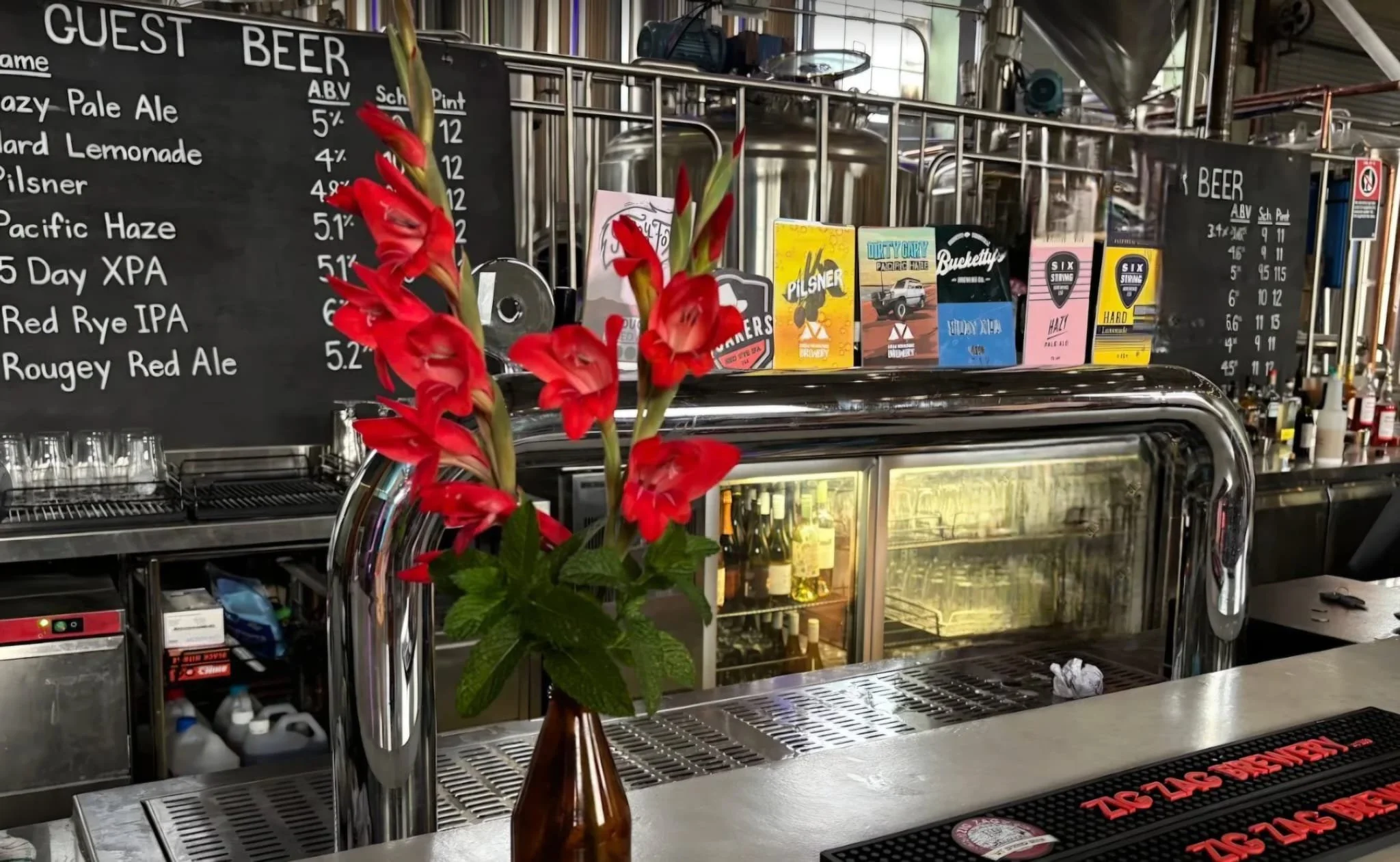 Red flowers in a brown vase on a bar counter with beer menu and brewery equipment in the background.