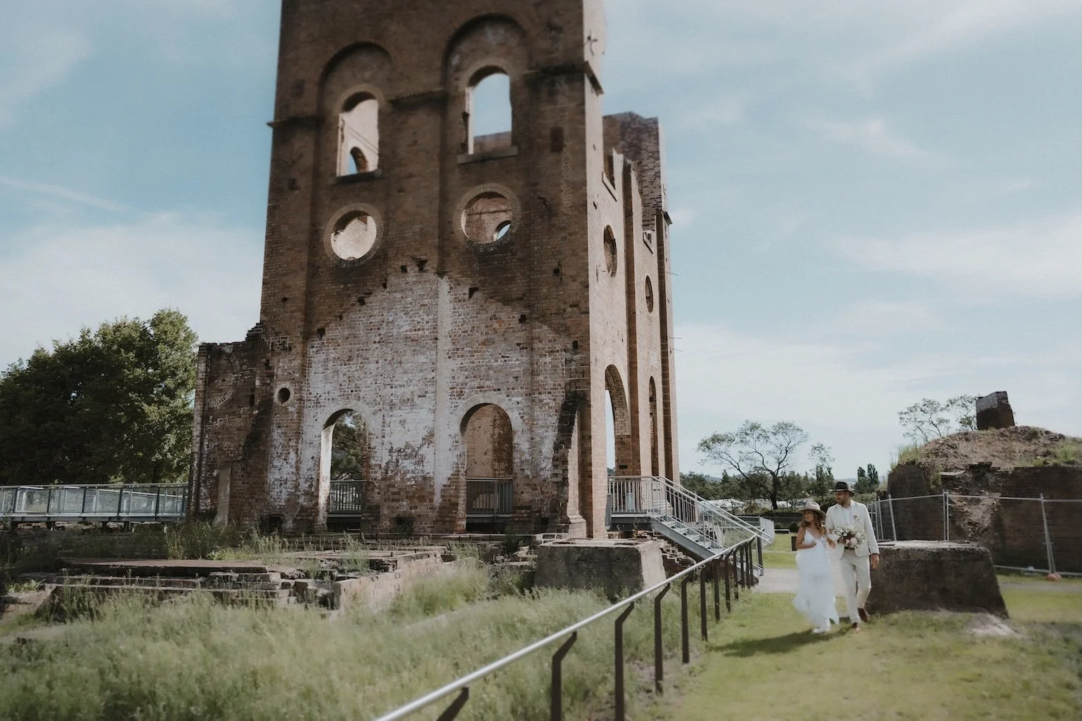 A bride and groom walking together in front of an ancient, partly ruined brick tower structure in a park, with trees and a cloudy sky in the background.