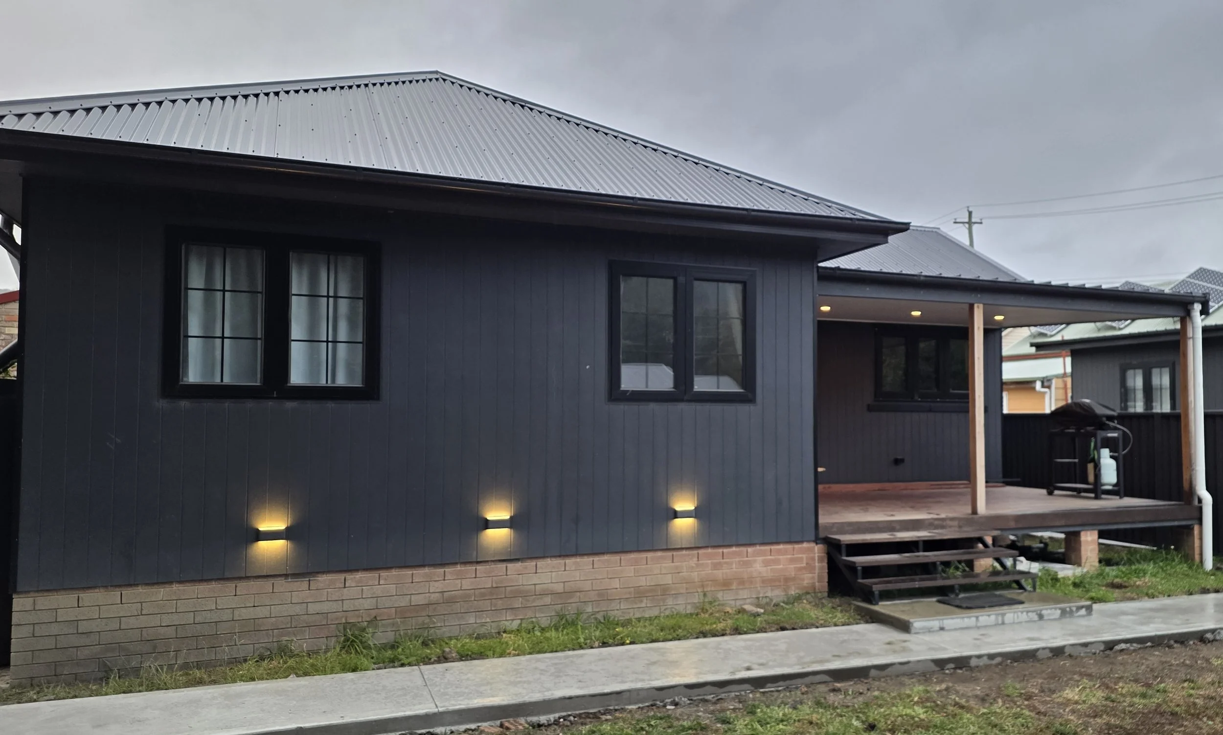 Modern house with dark exterior siding, brick foundation, illuminated outdoor lighting, and a metal roof, with steps leading to a porch and a barbecue grill nearby.