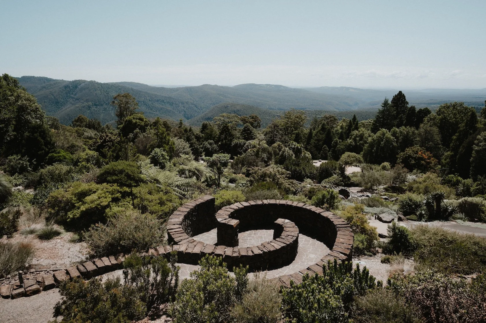 A scenic view of a lush green mountainside with a stone spiral structure in the foreground, under a clear blue sky.