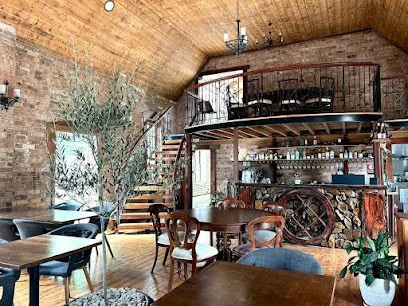 Interior of a rustic restaurant with a high wooden ceiling, exposed brick walls, and a mezzanine level. There are several tables and chairs, a large potted plant, and a bar with shelves of bottles in the background.