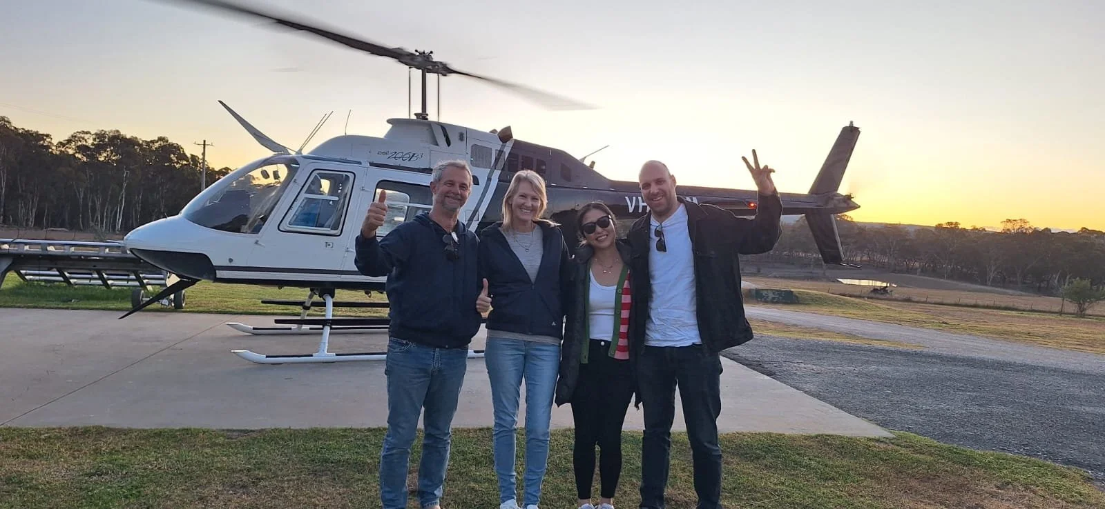 Four people standing in front of a helicopter at sunset, smiling and giving thumbs up and peace signs.