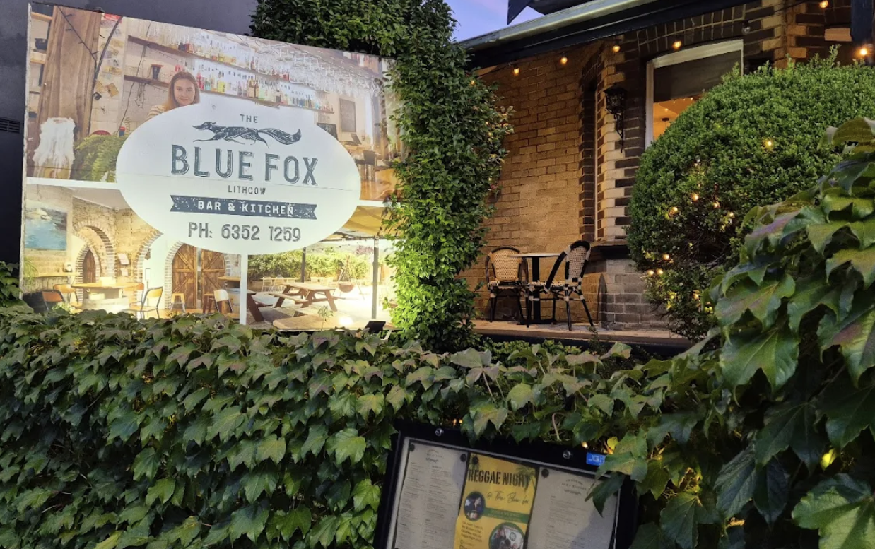 Sign for The Blue Fox Lithgow bar and kitchen with a phone number, located outside a brick building with outdoor seating, greenery, and string lights at dusk.