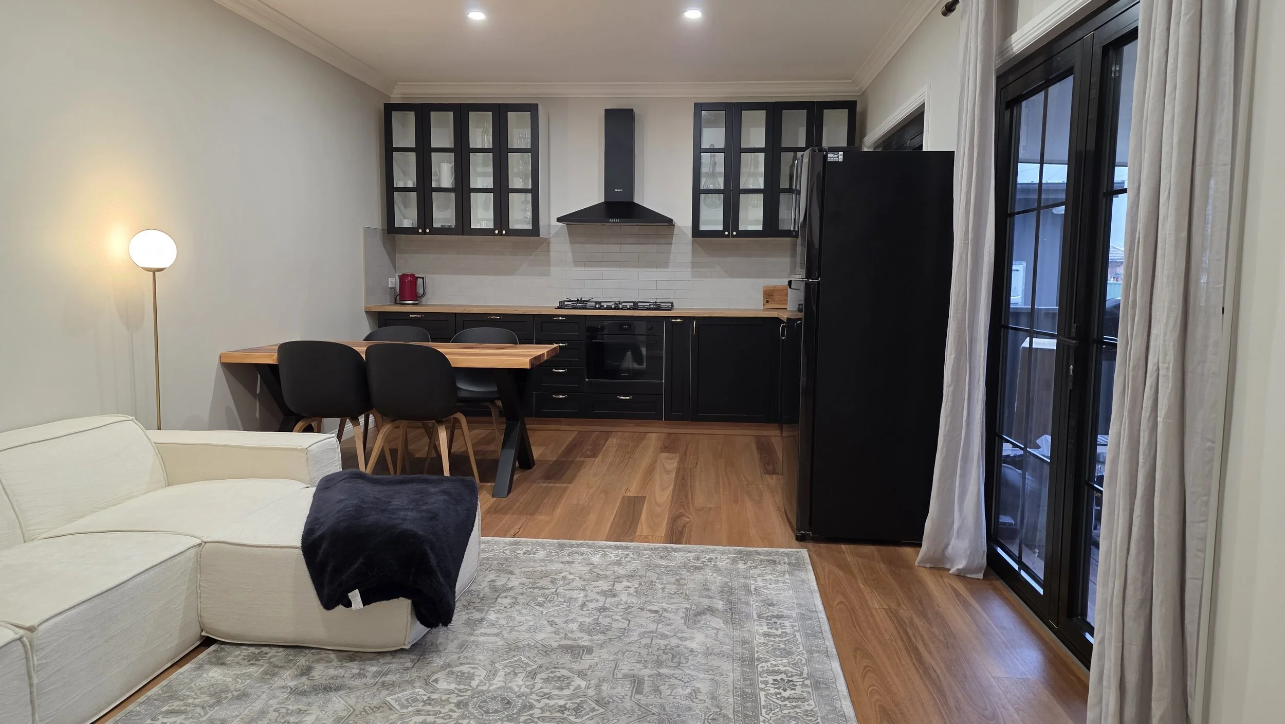 Interior view of a modern kitchen and living room with a cream sofa, black cabinetry, a wooden dining table with black chairs, and large windows with white curtains.