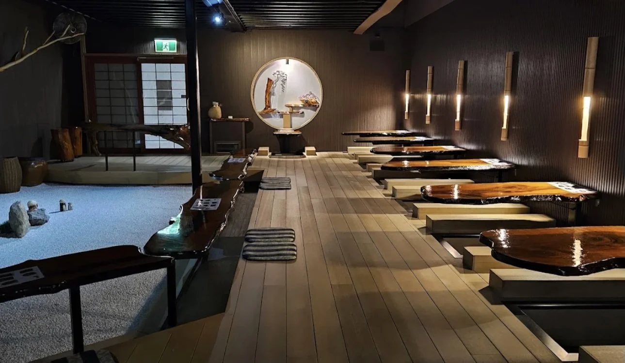 Interior of a Japanese restaurant with low wooden tables along the right wall, cushions for sitting, and a traditional decor with a circular window, bonsai, and small rocks.