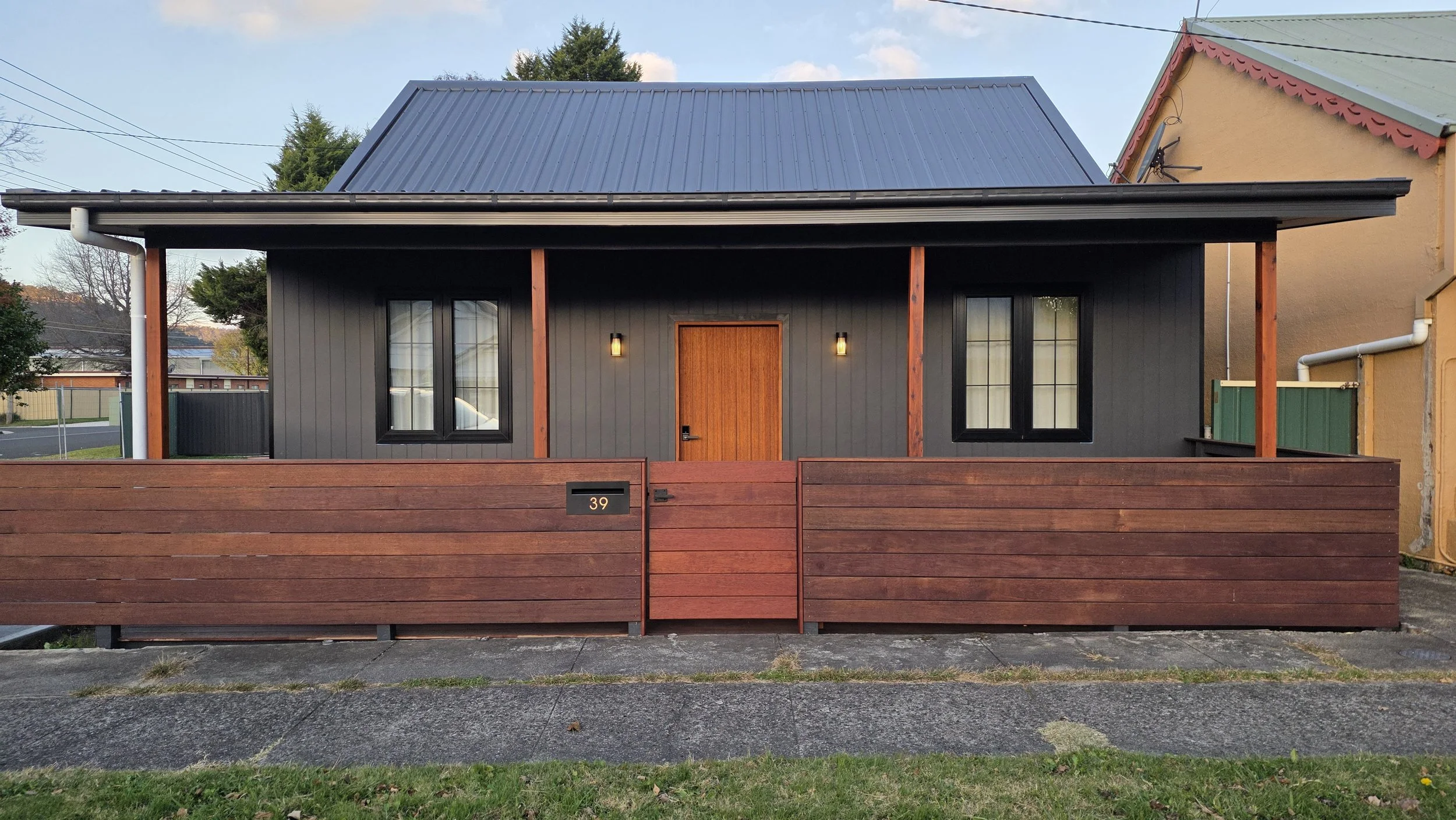 Front view of a modern house with black siding, two windows, a wooden front door, exterior wall-mounted lights, and a horizontal wooden fence with house number 39.