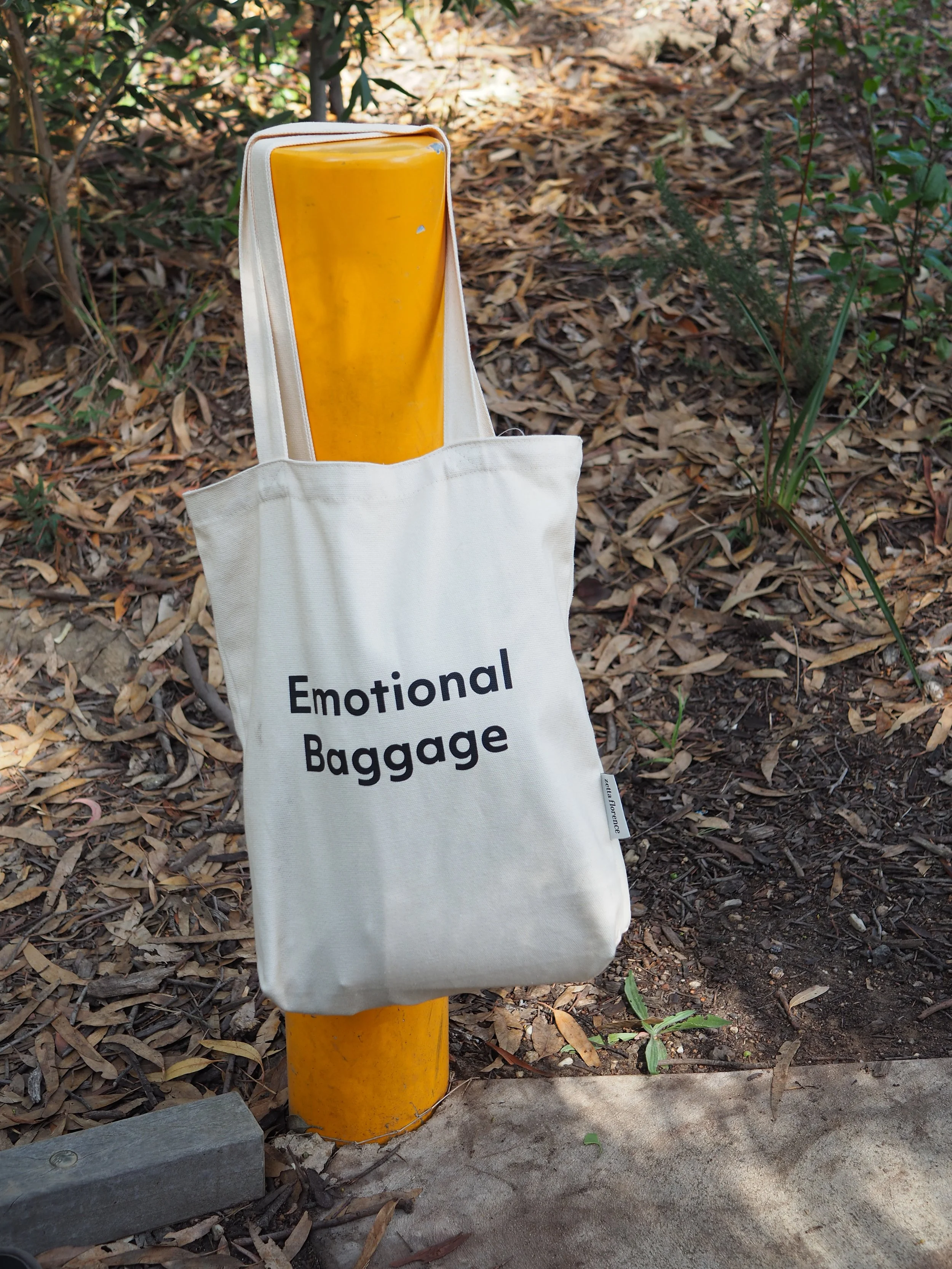 A canvas tote bag printed with the words Emotional Baggage, leaning against a yellow bollard in an outdoor setting