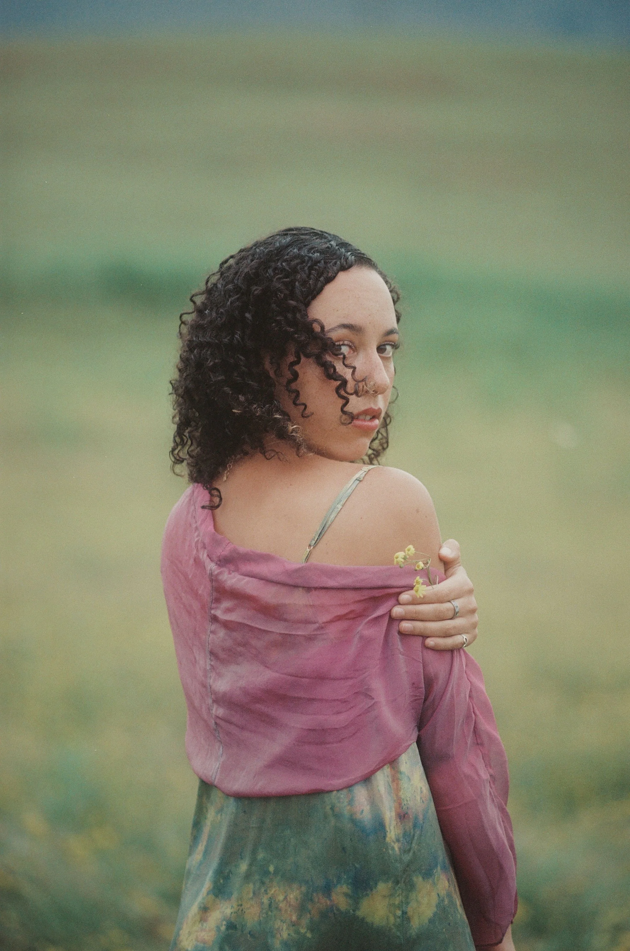 A woman with curly dark hair looks over her shoulder at the camera, holding flowers and wearing a pink off-shoulder top against a blurred green and blue background.
