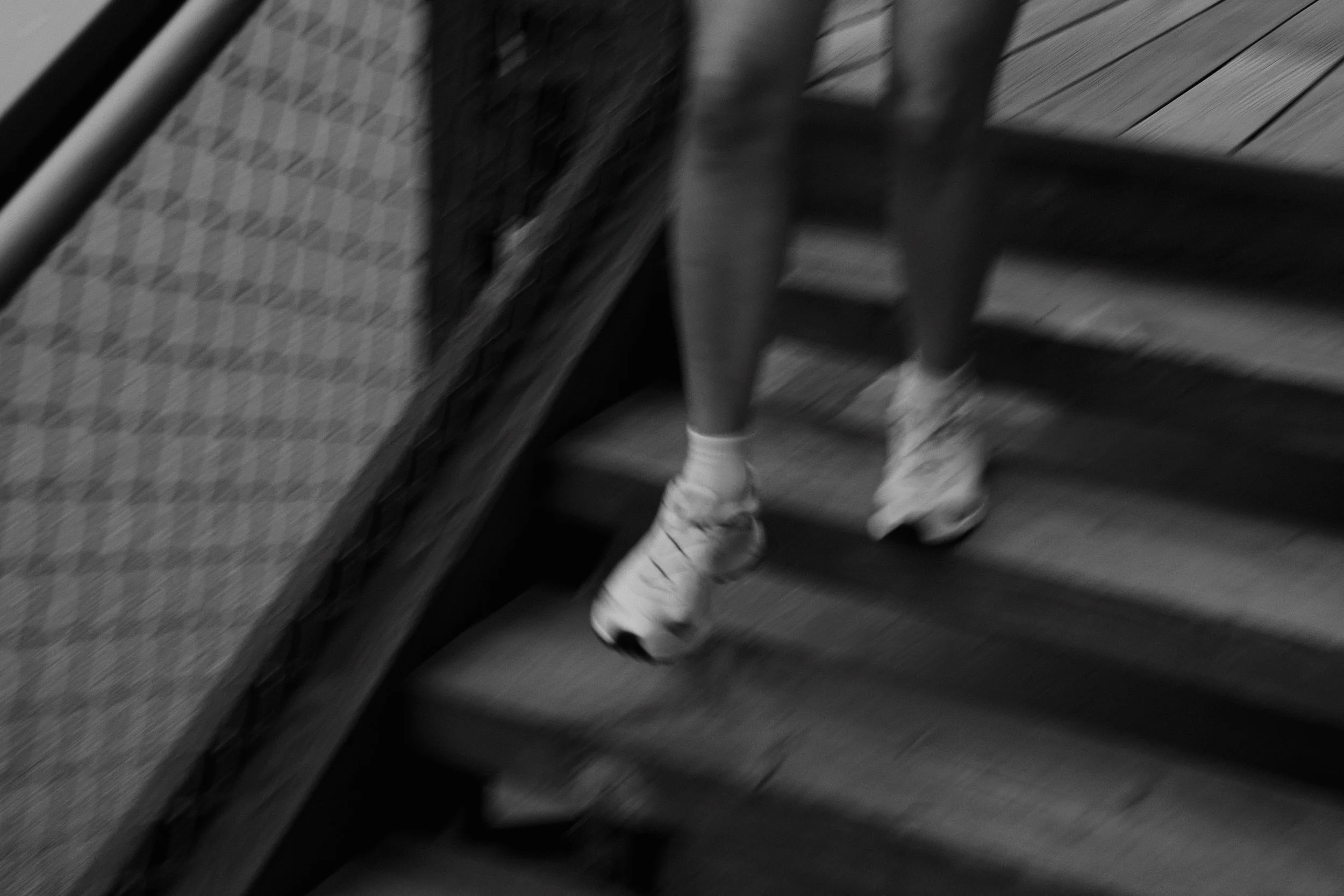 Blurry black and white photo of a person's legs and feet wearing striped socks, standing on a staircase near a railing and a wall