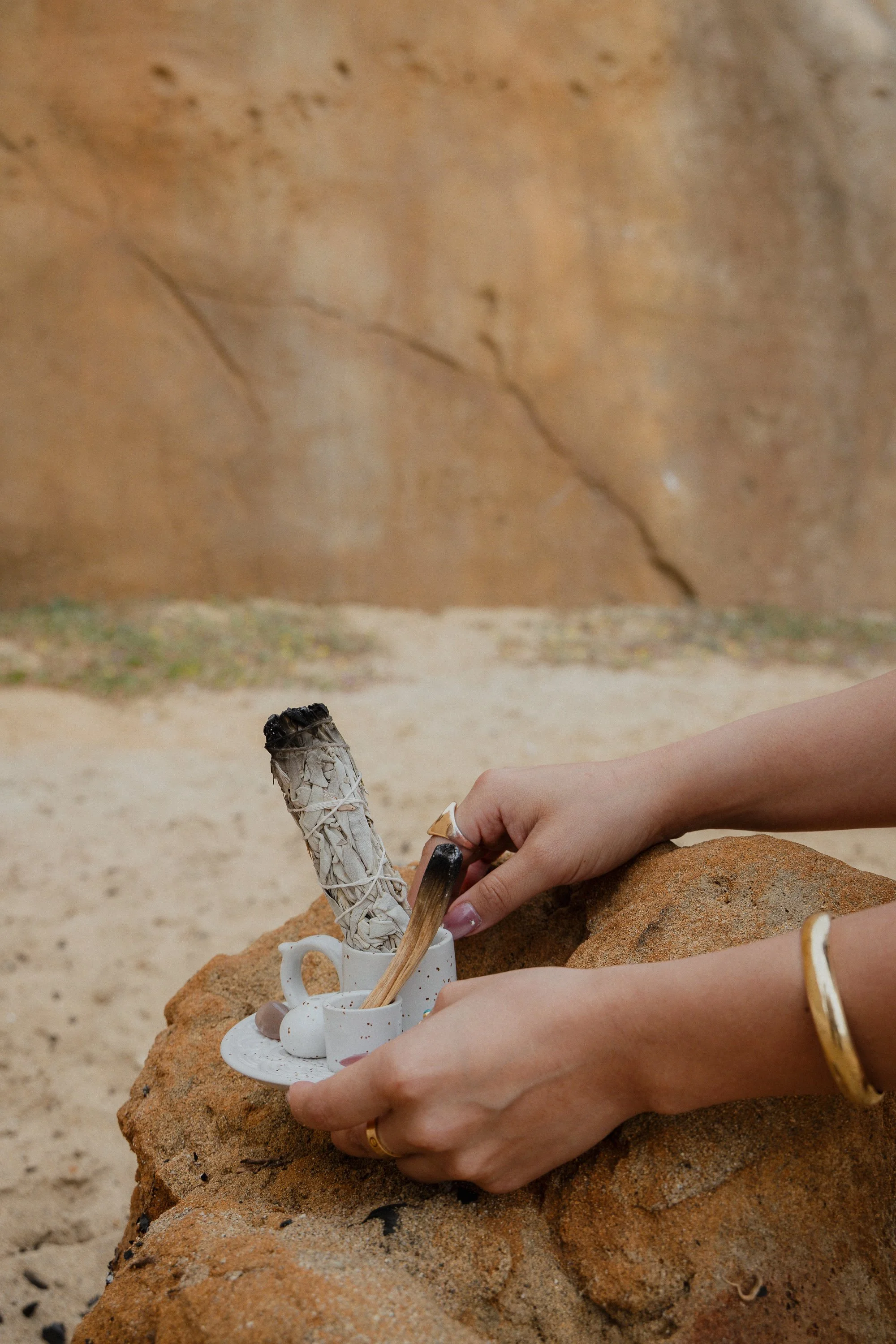 A person's hands arranging items in a white mug with a small white teacup, a small white jar, and a wooden stick, on a large rock on a sandy surface with a blurred beige wall in the background.