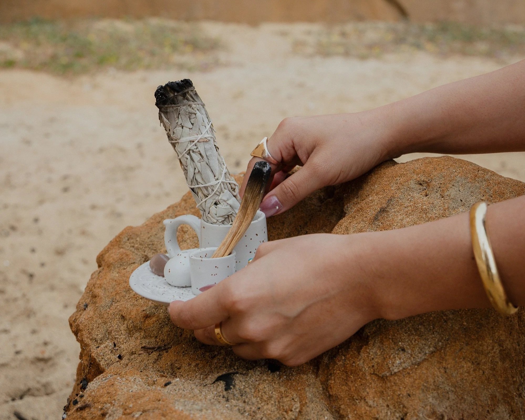 A person's hand holding a white ceramic mug with a sand-colored ring on the finger, placed on a rust-colored rock with a small white ceramic object, against a blurry sandy background.