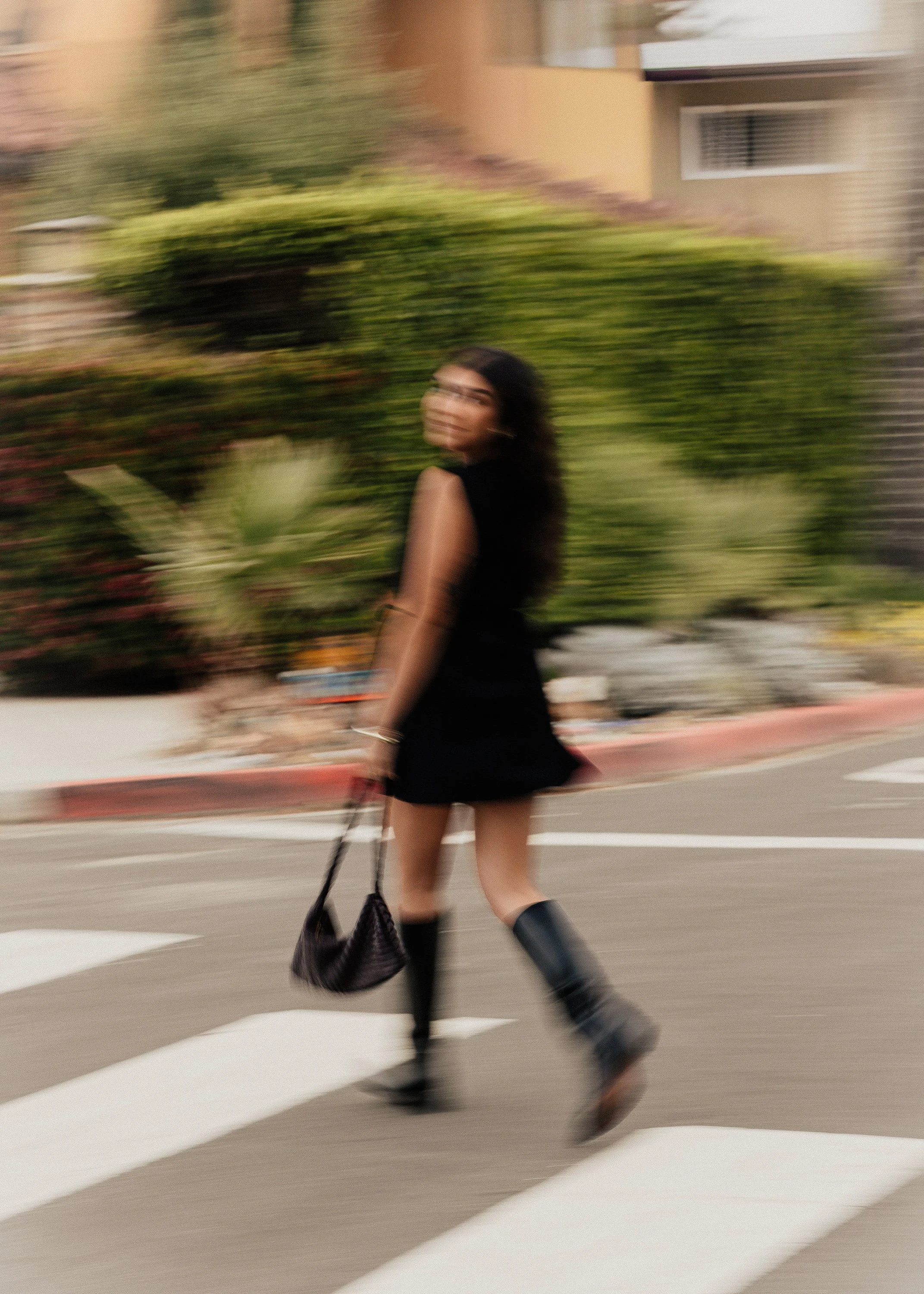 A woman walking across a pedestrian crosswalk, carrying a purse and wearing black boots and a black outfit, with a blurred background of greenery and buildings.