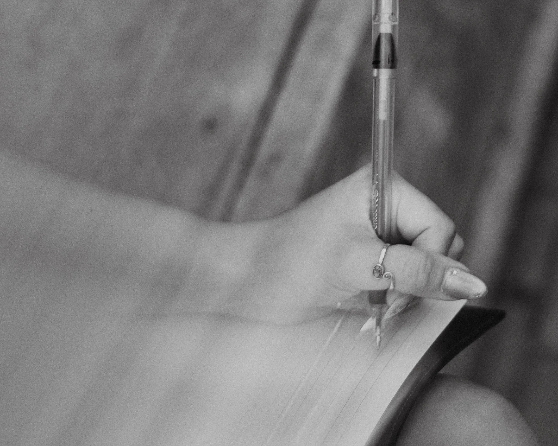 Close-up of a person writing with a pen on paper. The person's hand is visible, wearing a bracelet, resting on paper, with the pen held in the other hand. The background is blurred.