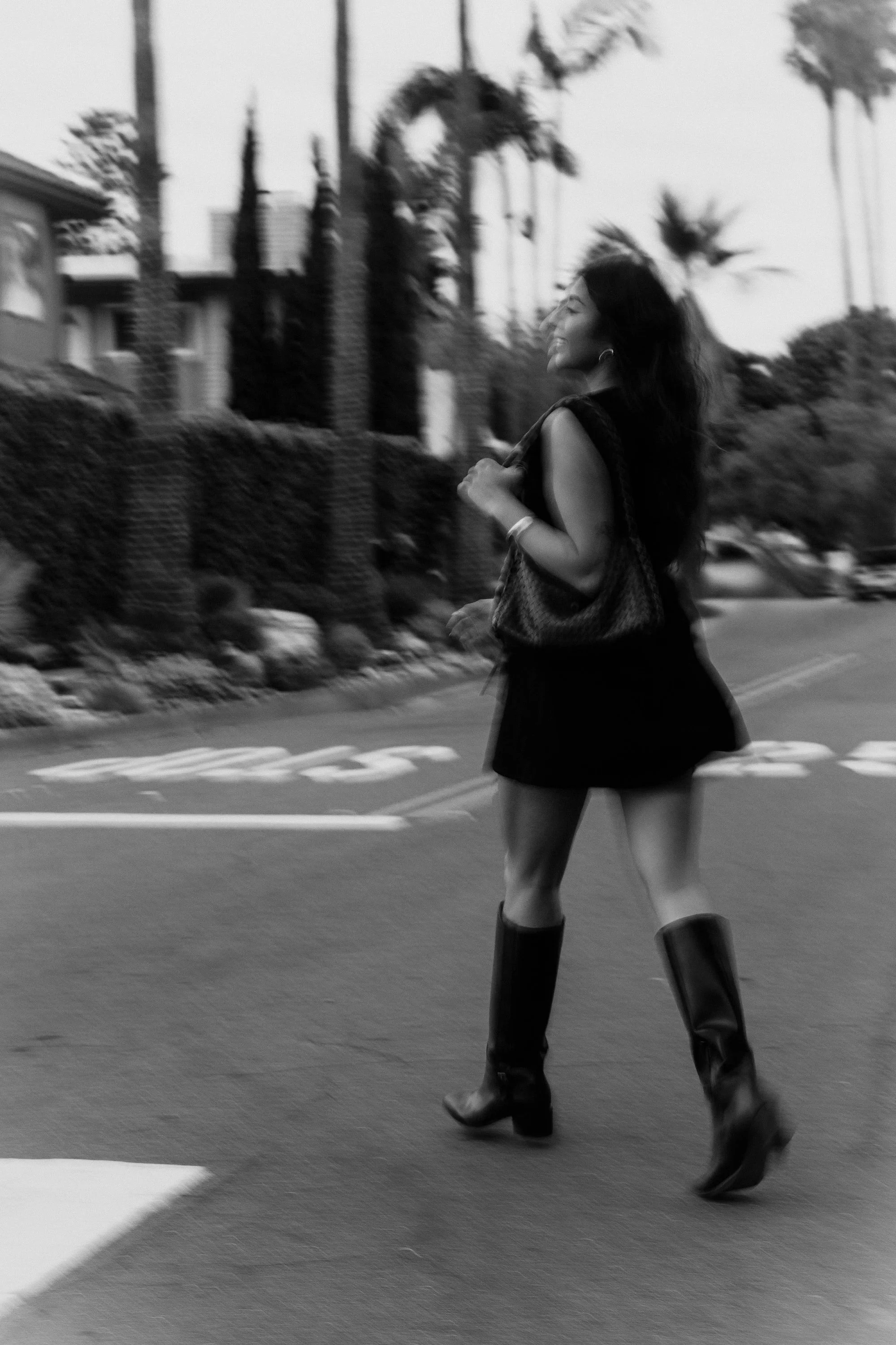 A woman with long hair smiling and walking on a street, wearing a sleeveless top, a short skirt, and knee-high boots, with palm trees and buildings in the background.