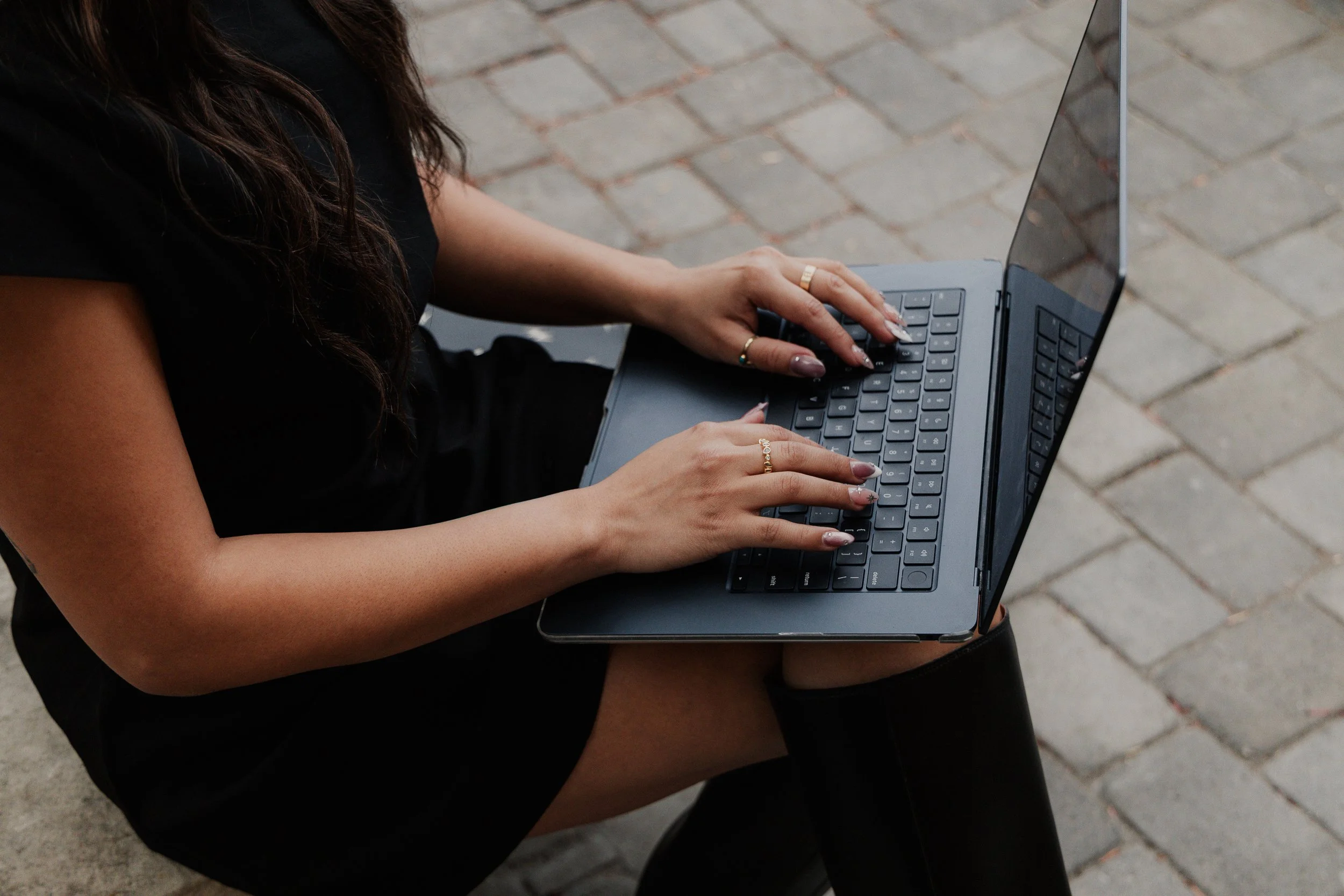 A woman with long dark hair is sitting outdoors on a black chair, using a gray laptop placed on her lap. She is wearing a black top and appears to be working or browsing, with a brick pavement visible in the background.