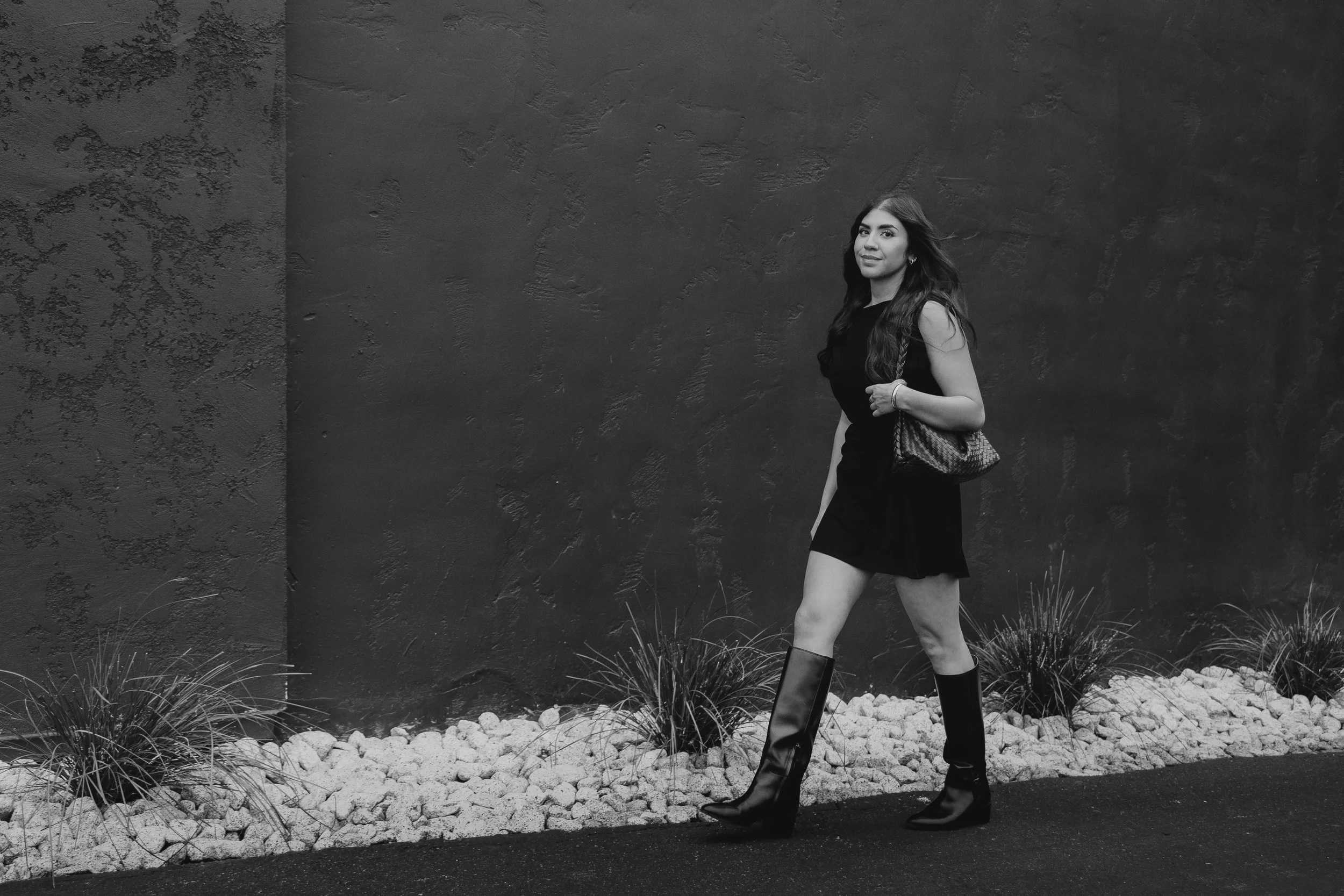 A woman walking outdoors against a textured wall, wearing a black dress, knee-high boots, and carrying a patterned handbag.