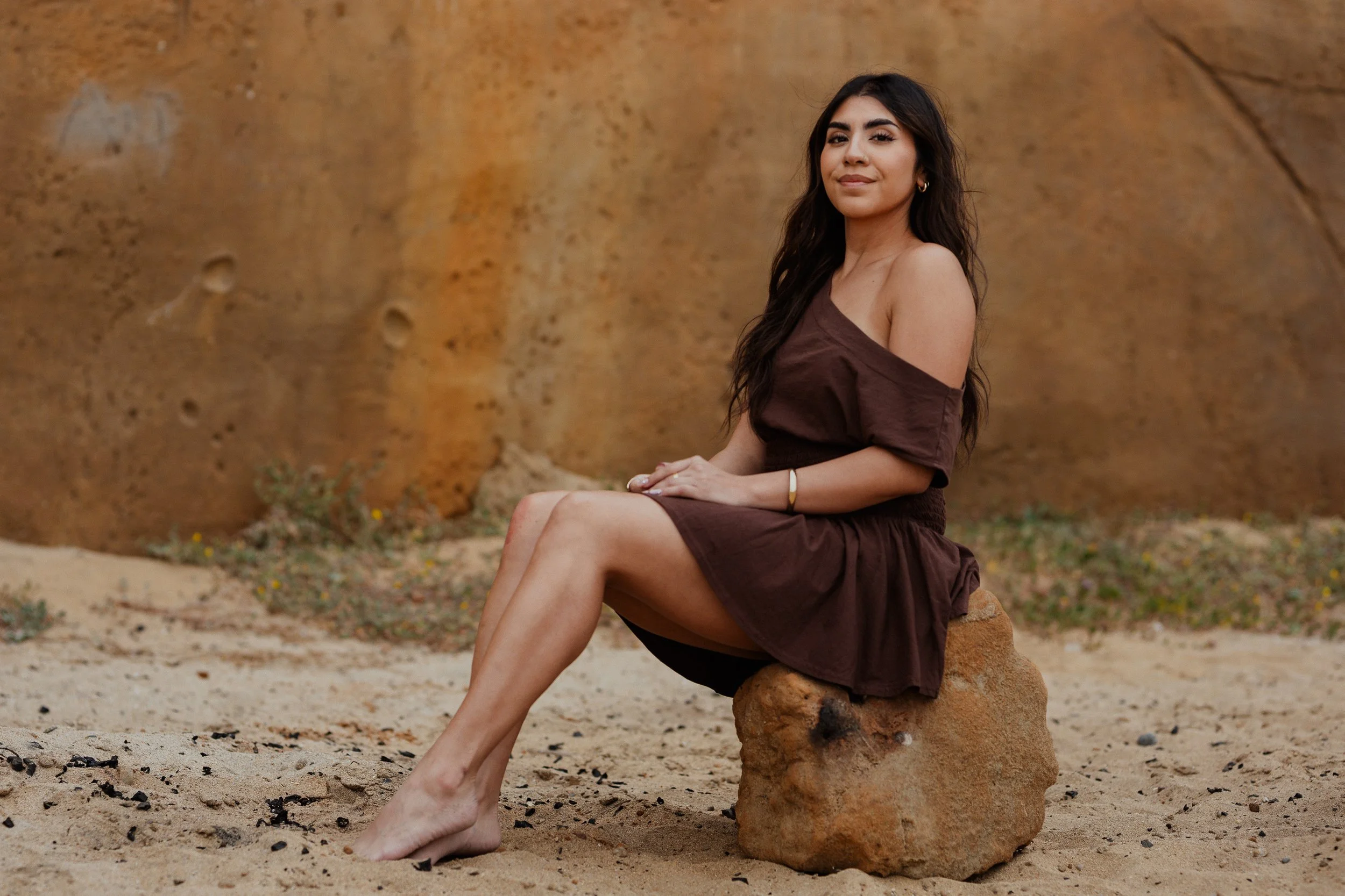 A woman with long dark hair sitting on a rock in a desert, wearing a brown off-shoulder dress, with a large rock wall in the background.