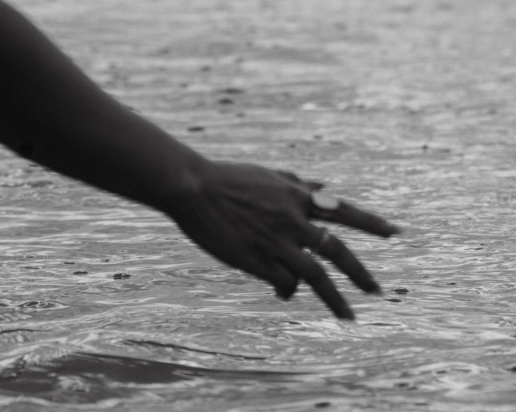 Close-up of a person's hand near the water on a sandy beach, with the sea in the background, in black and white.