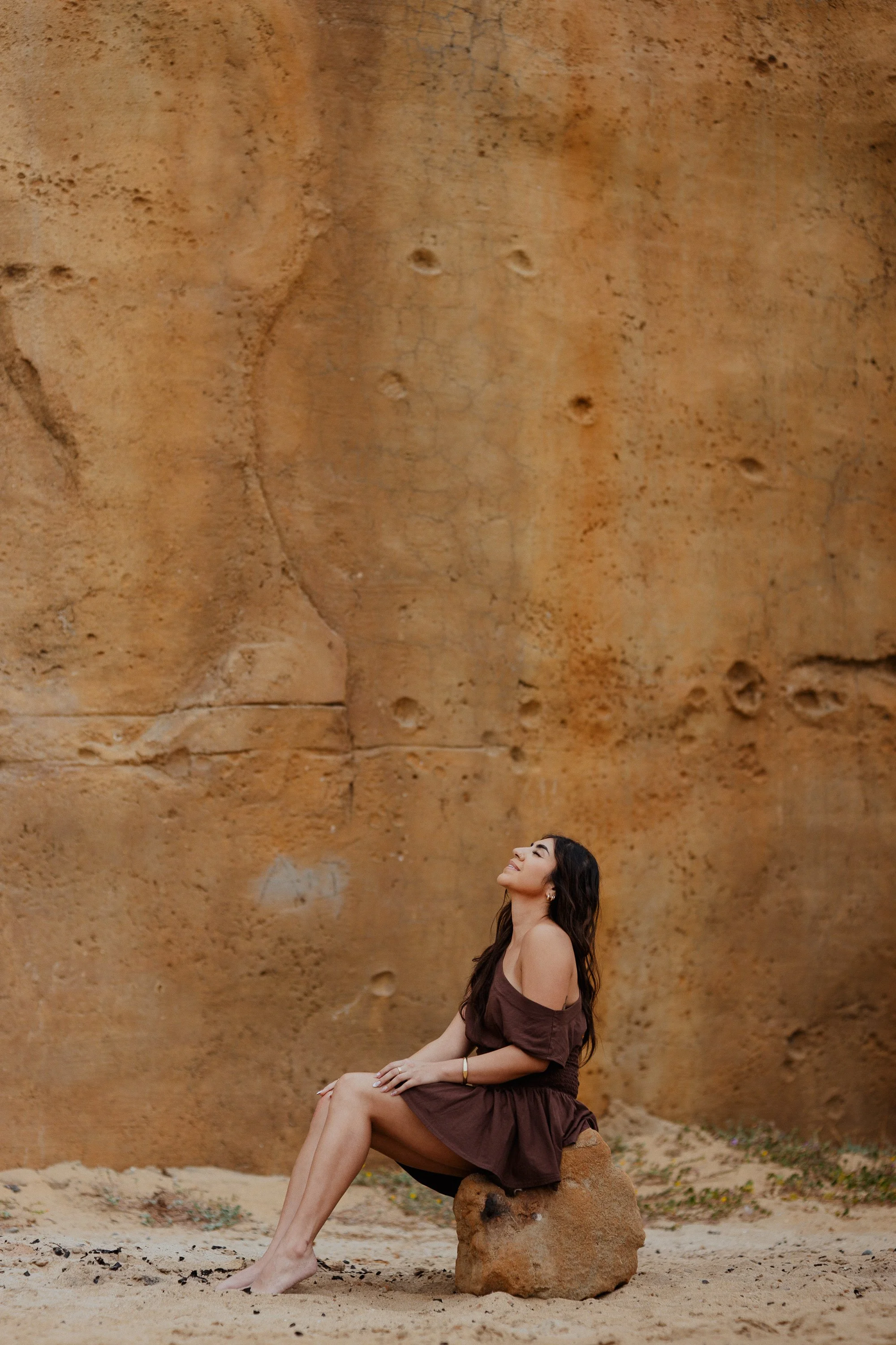 A woman sitting on a rock with her legs crossed, smiling and looking up, against a large, textured, orange-brown rock formation that resembles a cliff or canyon wall.