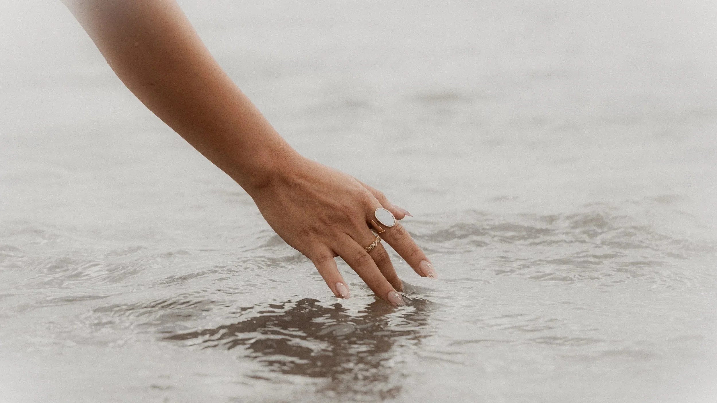 A person's hand touches the water on a sandy beach, with the sky and water in the background.