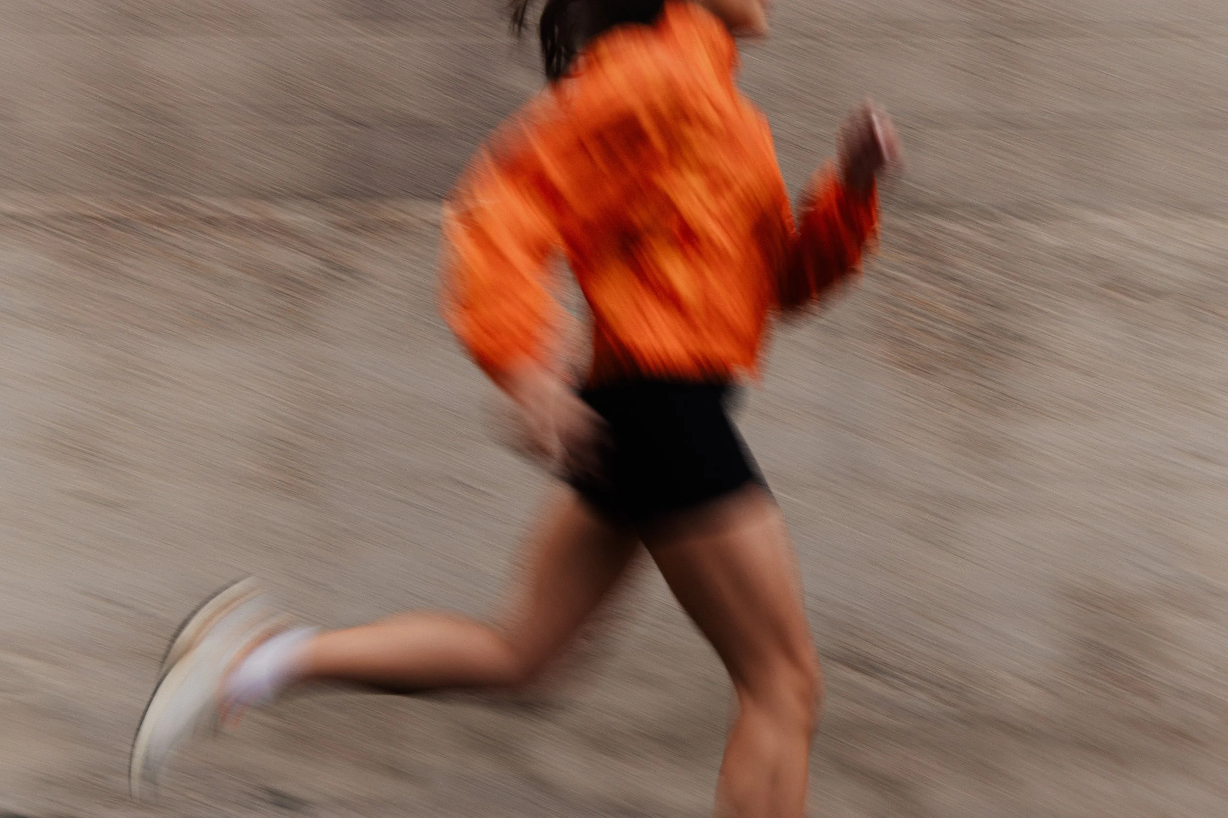 Blurry motion photo of a person running or jogging on a gravel or concrete surface, wearing an orange top, black shorts, and white shoes.
