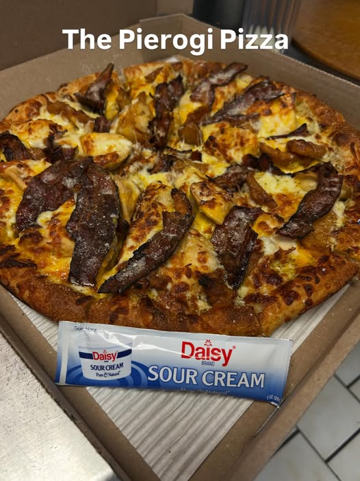 A pizza with bacon and cheese in a cardboard box, with a packet of Daisy sour cream in front. The caption reads 'The Pierogi Pizza'.
