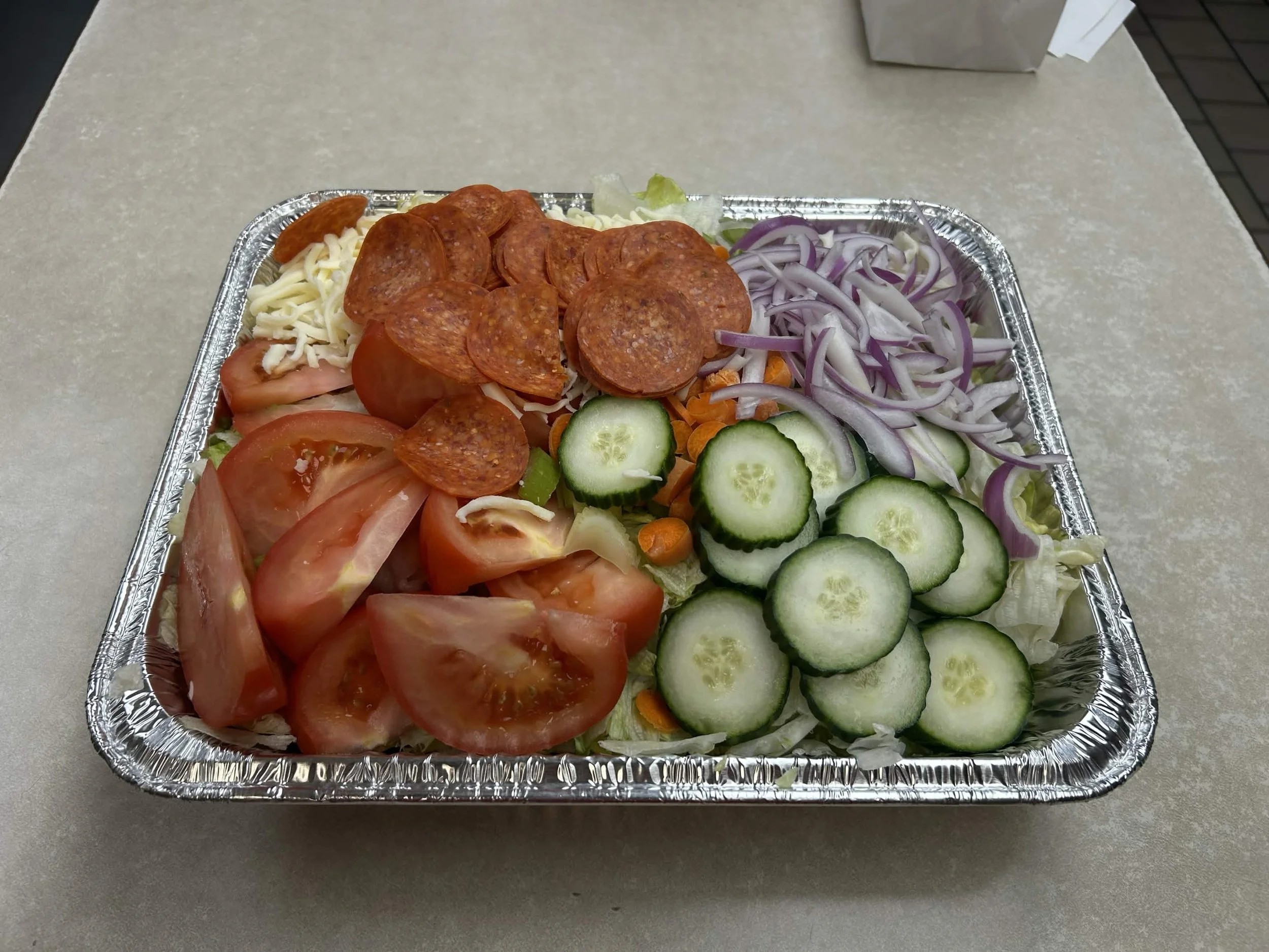 A foil tray filled with sliced tomatoes, cucumbers, shredded lettuce, sliced onions, shredded cheese, sliced pepperoni, shredded carrots, and lettuce.
