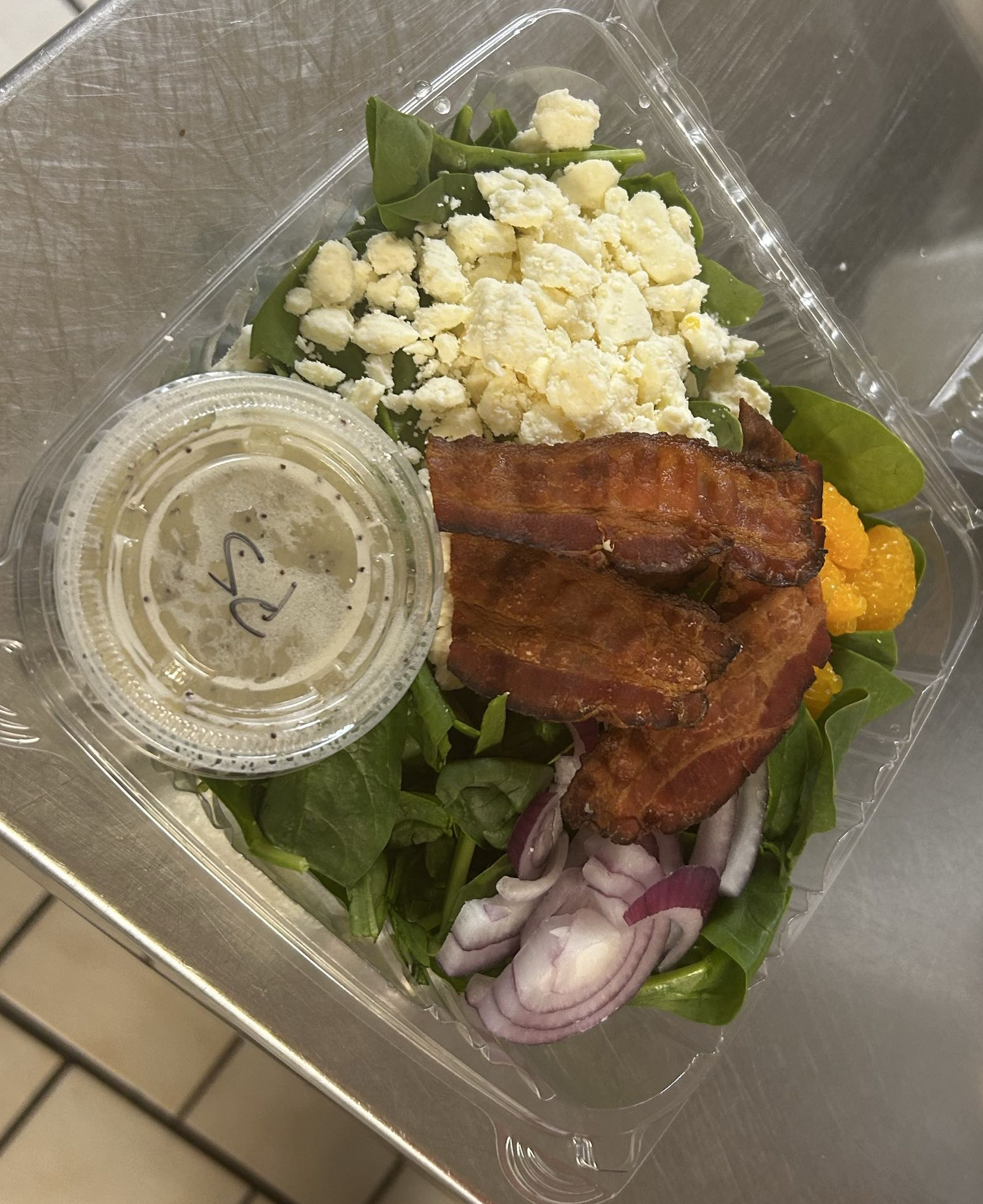 Salad with spinach, shredded cheese, bacon strips, sliced onions, shredded cheddar cheese, and a small container of salad dressing.
