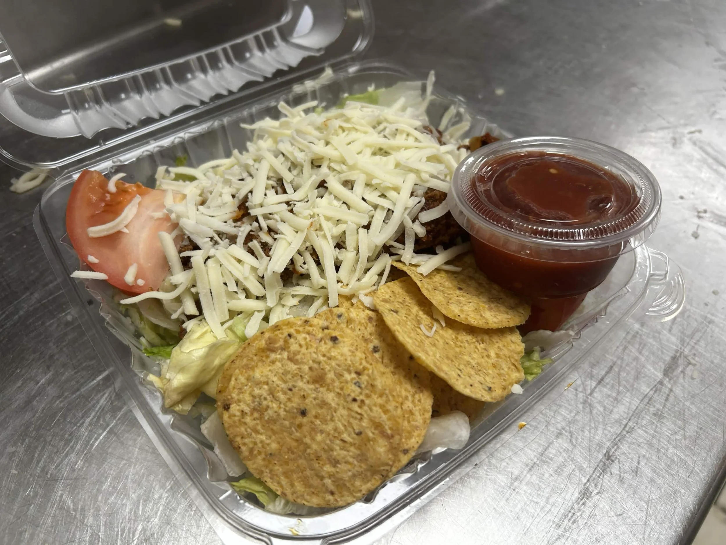 A takeout container holding a taco salad with shredded cheese, lettuce, tomato, taco chips, and a side of salsa sauce.