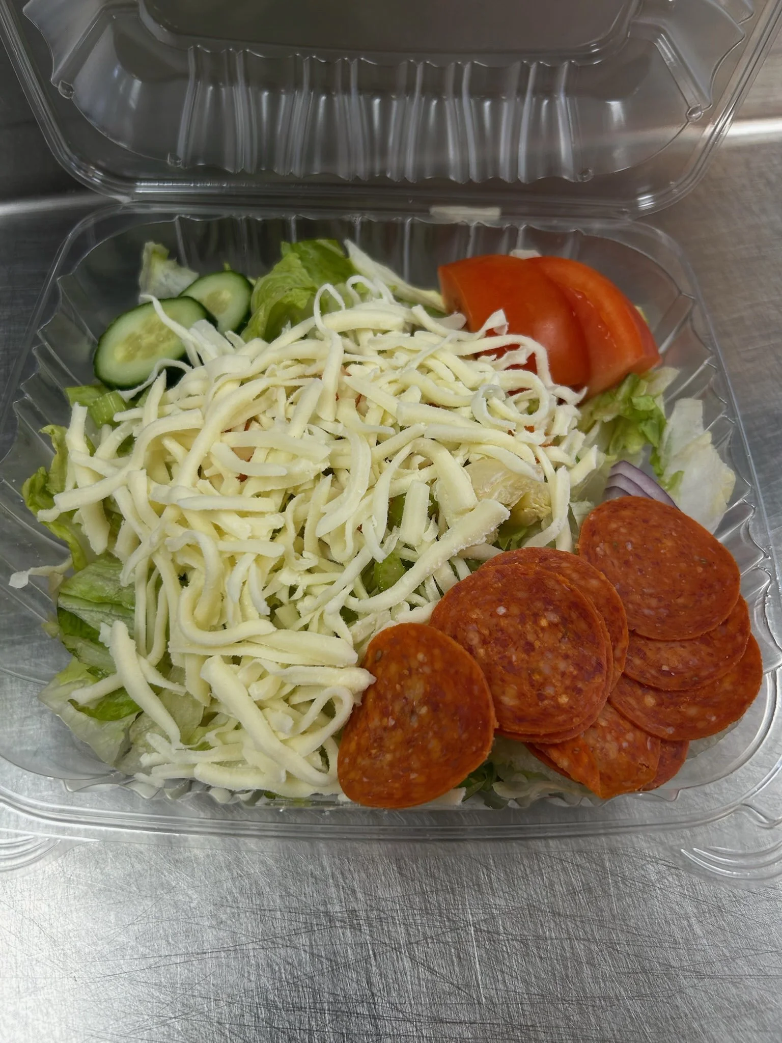 Salad with shredded cheese, sliced cucumber, tomato wedges, lettuce, and pepperoni slices in a plastic container on a metallic surface.