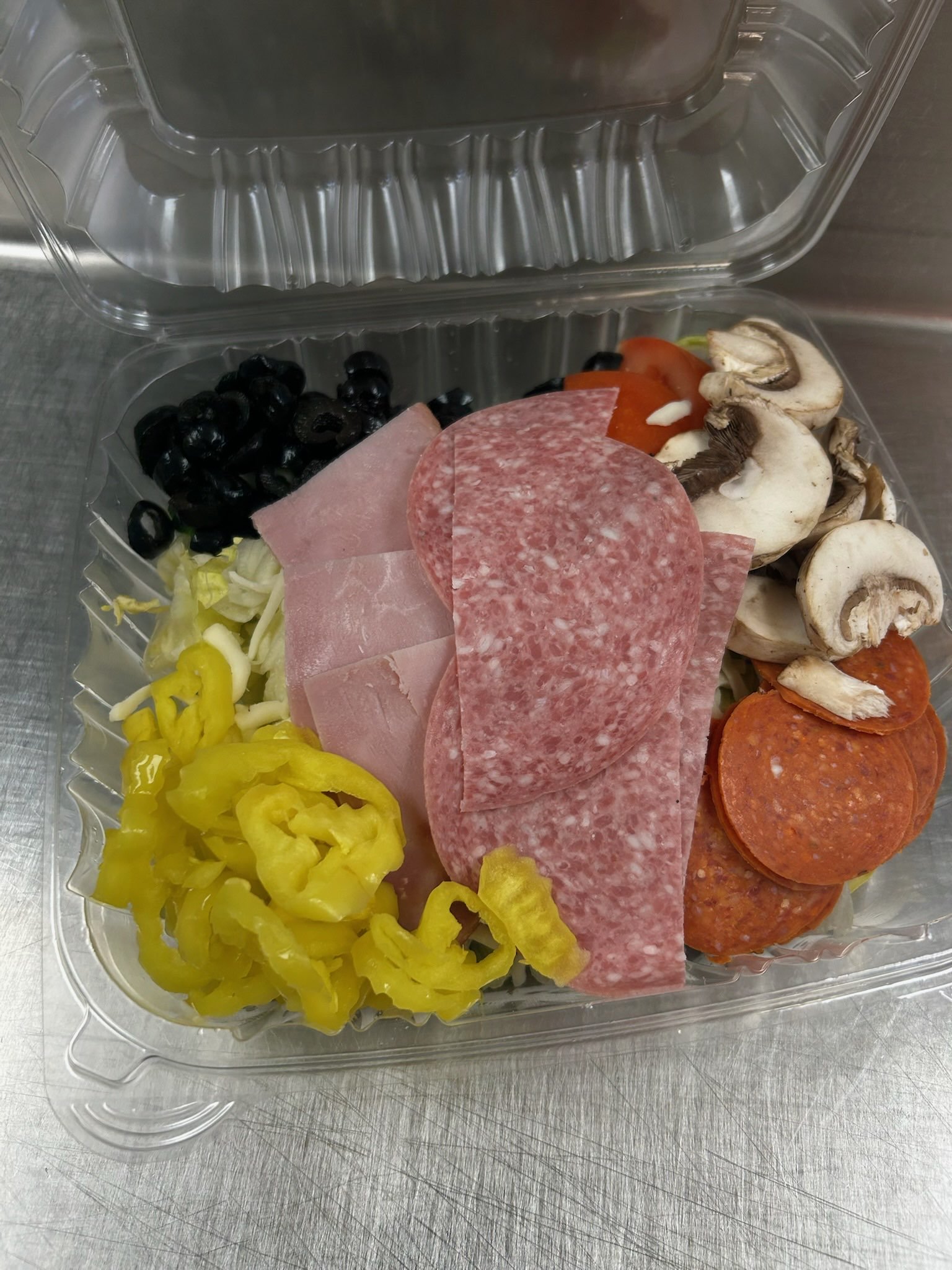 Open plastic container with yellow peppers, black olives, sliced ham, salami, mushrooms, tomato slices, and shredded lettuce, all on a stainless steel surface.