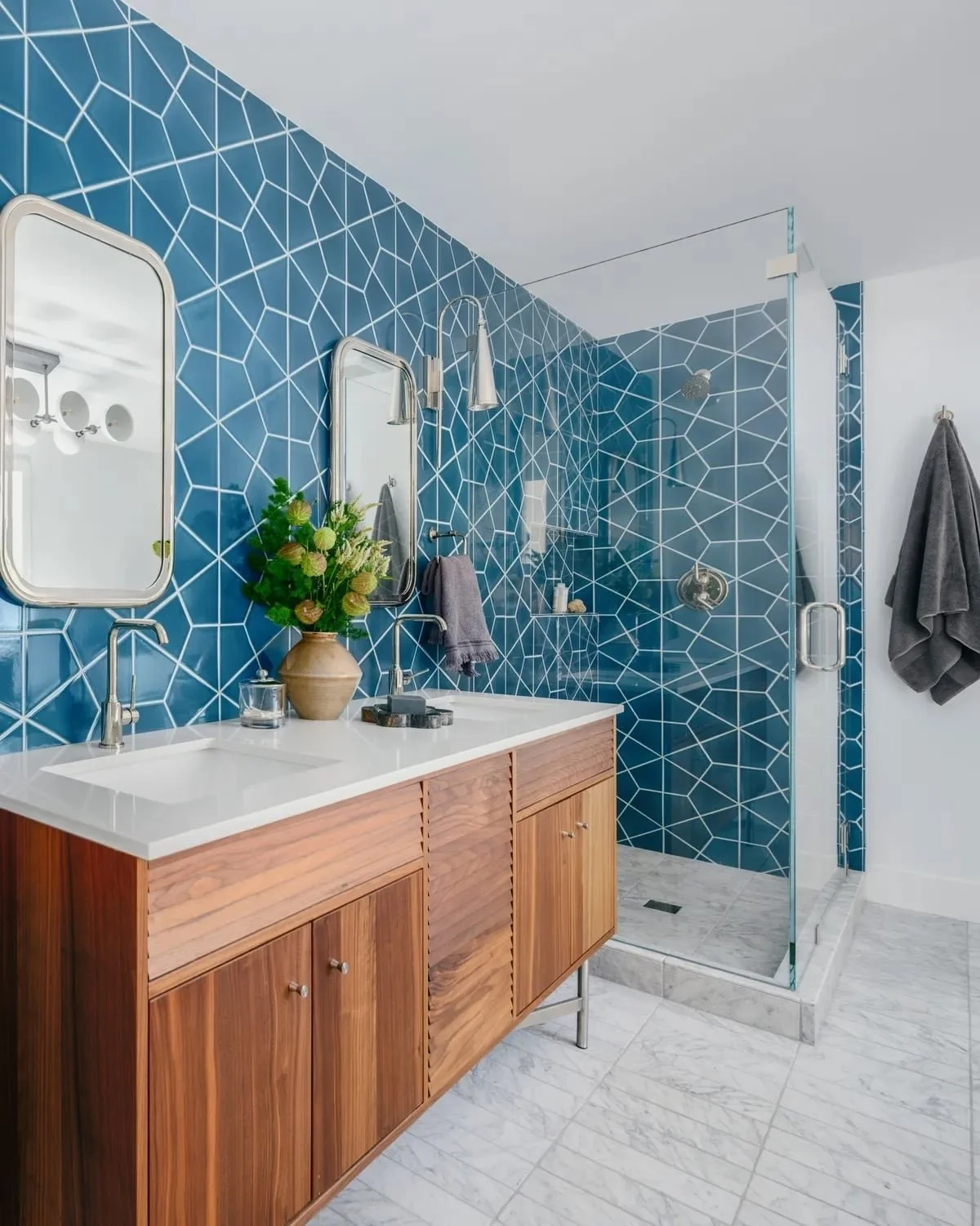 A modern bathroom featuring a blue geometric tile wall, a wooden vanity with a white countertop, twin oval mirrors, and a glass-enclosed shower with matching blue tiles.