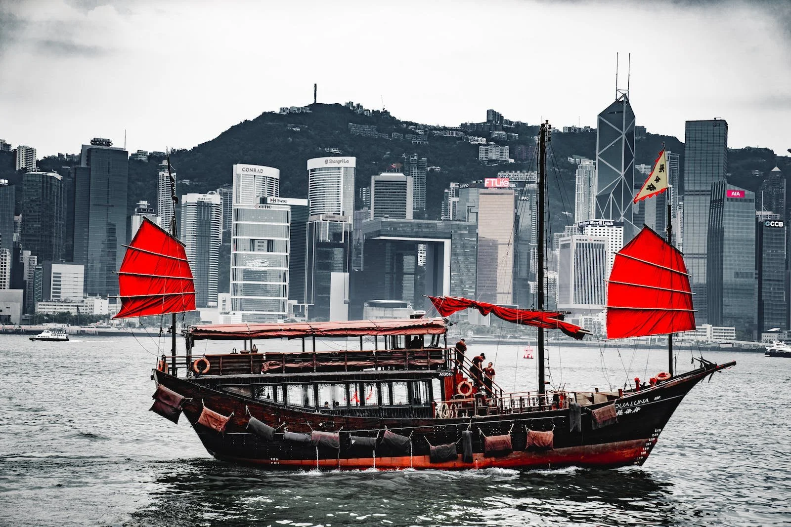 A traditional boat with red sails and black hull sailing on water, with a modern city skyline and mountain in the background.