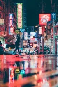 Nighttime city street scene with neon signs, pedestrians, and reflections on wet pavement.