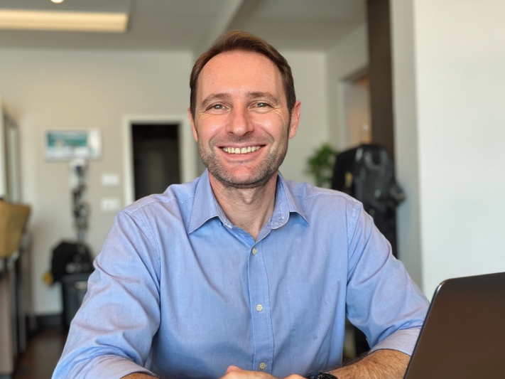 Smiling man in a blue shirt sitting at a table in a modern indoor setting with a laptop in front of him.