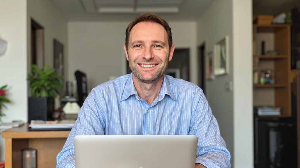 A man sitting at a desk smiling at the camera with a laptop in front of him, in a well-lit room with bookshelves and plants in the background.