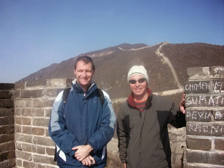 Two men standing on the Great Wall of China, with mountains in the background and clear blue sky, smiling at the camera.