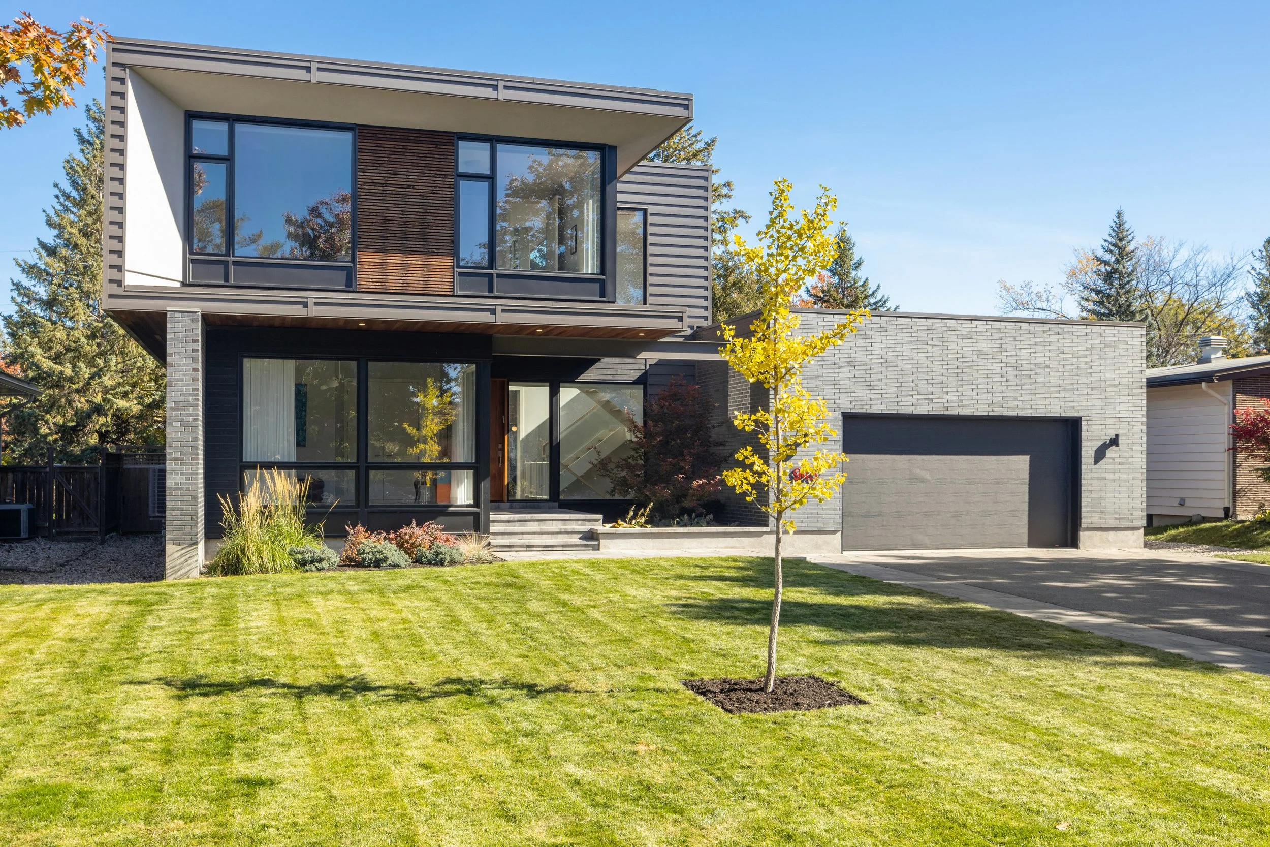 Modern two-story house with large windows, a gray exterior, and a landscaped yard with a small tree in the foreground.