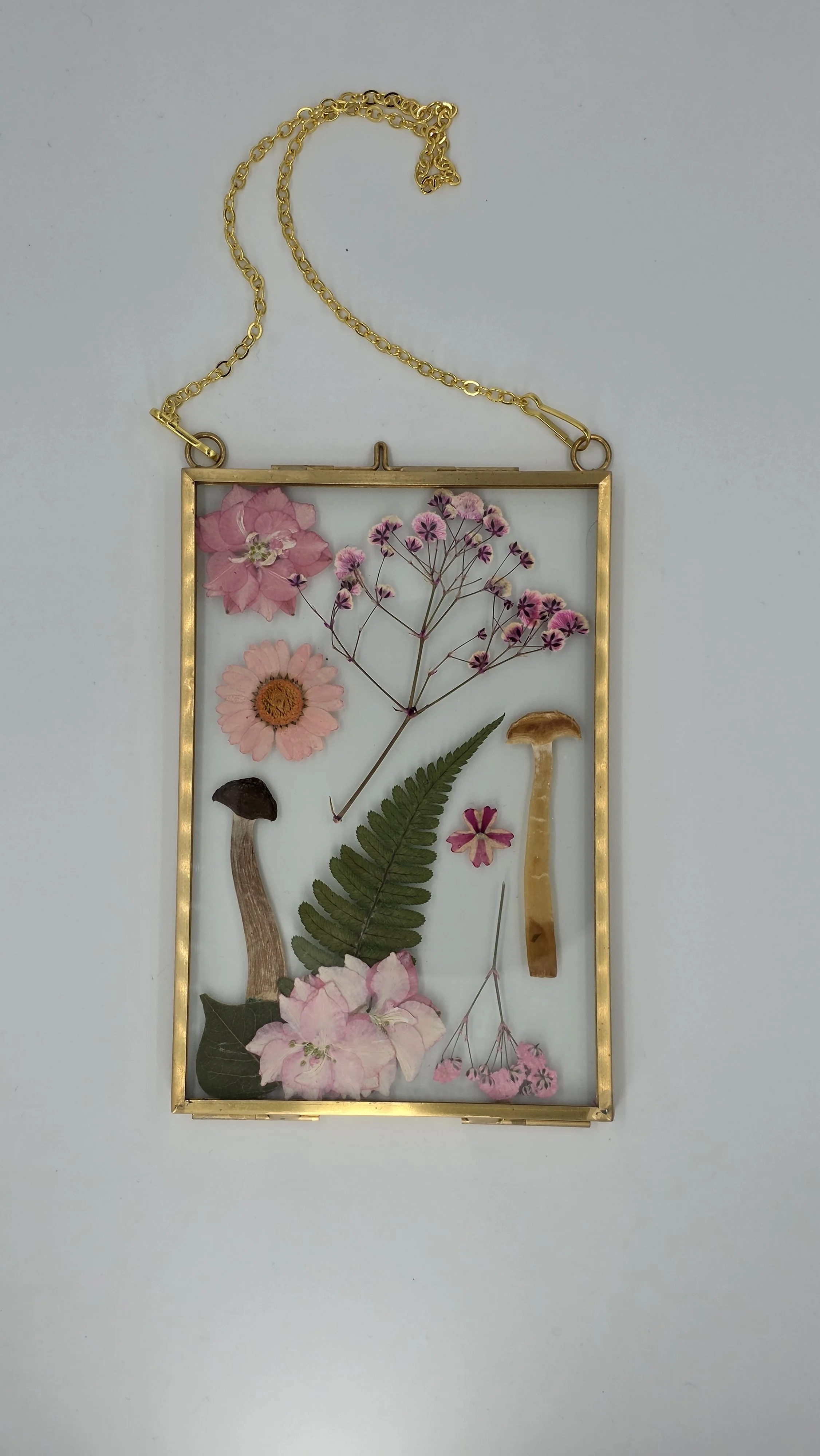 A gold frame with dried pink and purple flowers, a green fern, and two small mushroom-shaped objects inside, hanging from a gold chain.