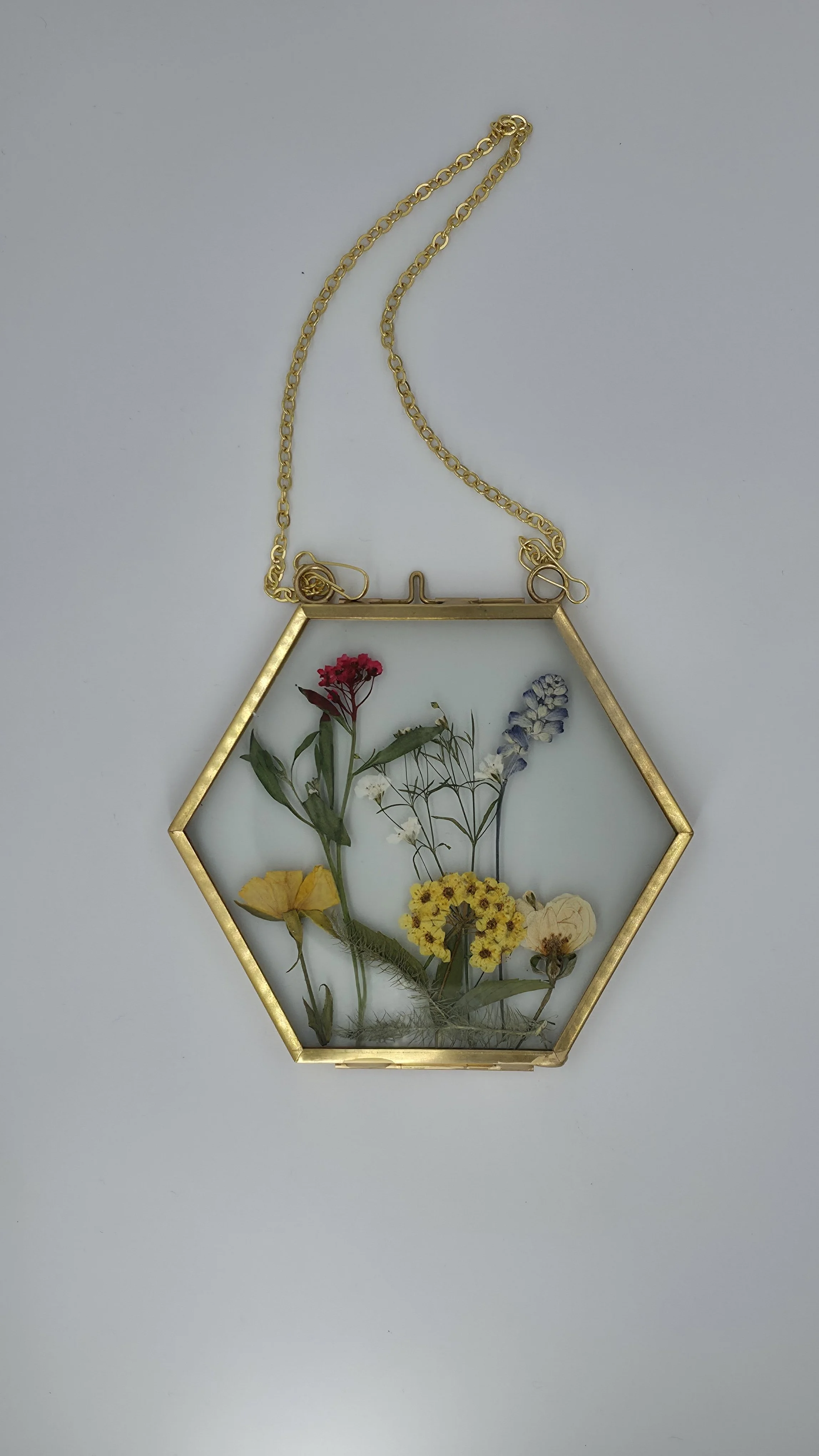 A gold hexagon-shaped frame with pressed flowers inside, hanging from a gold chain on a light gray background.