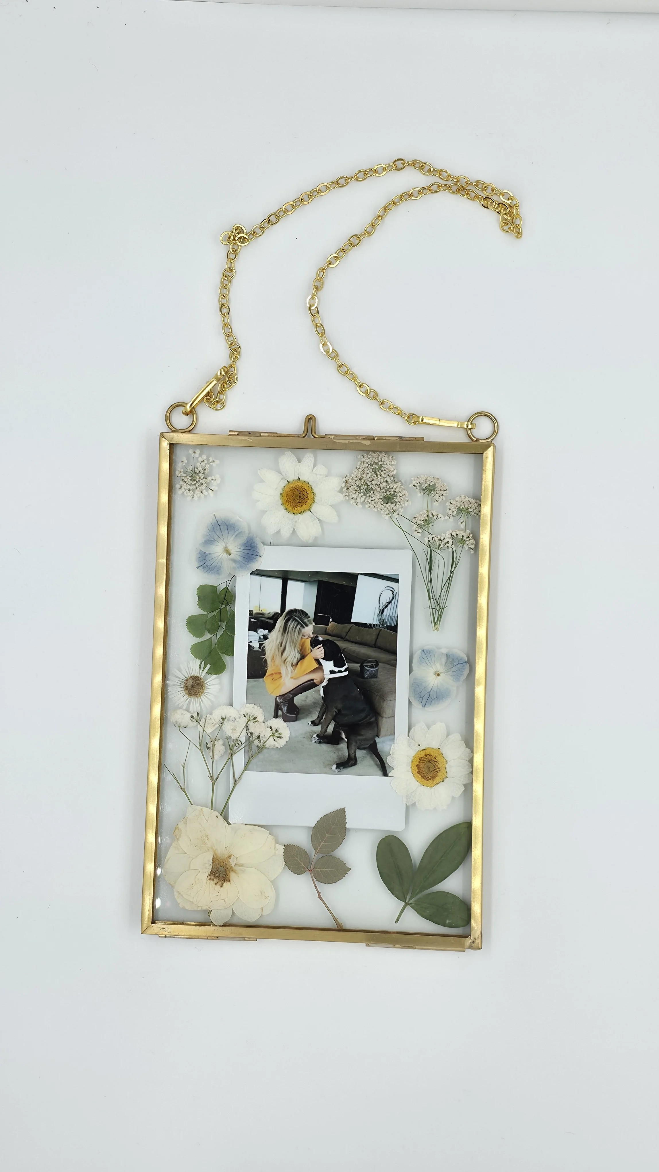 A decorative picture frame with a gold border hanging from a gold chain, filled with pressed flowers and a photo of a woman hugging a dog inside.