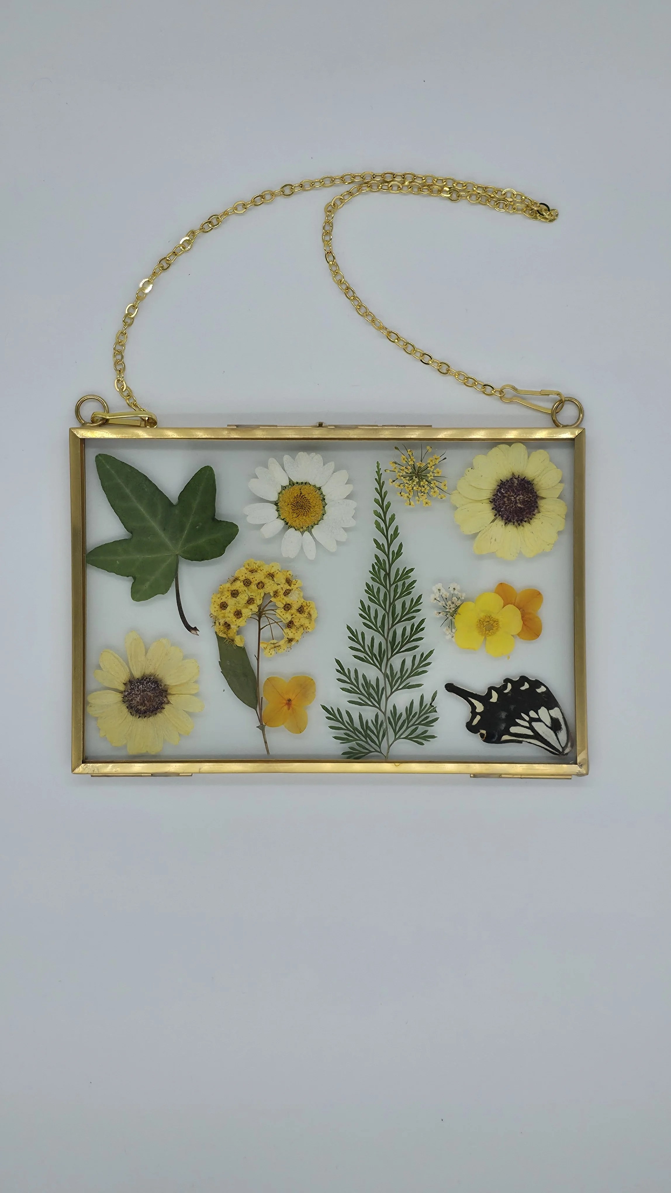 A glass frame display with pressed flowers and a butterfly silhouette inside, hanging from a gold chain on a white background.