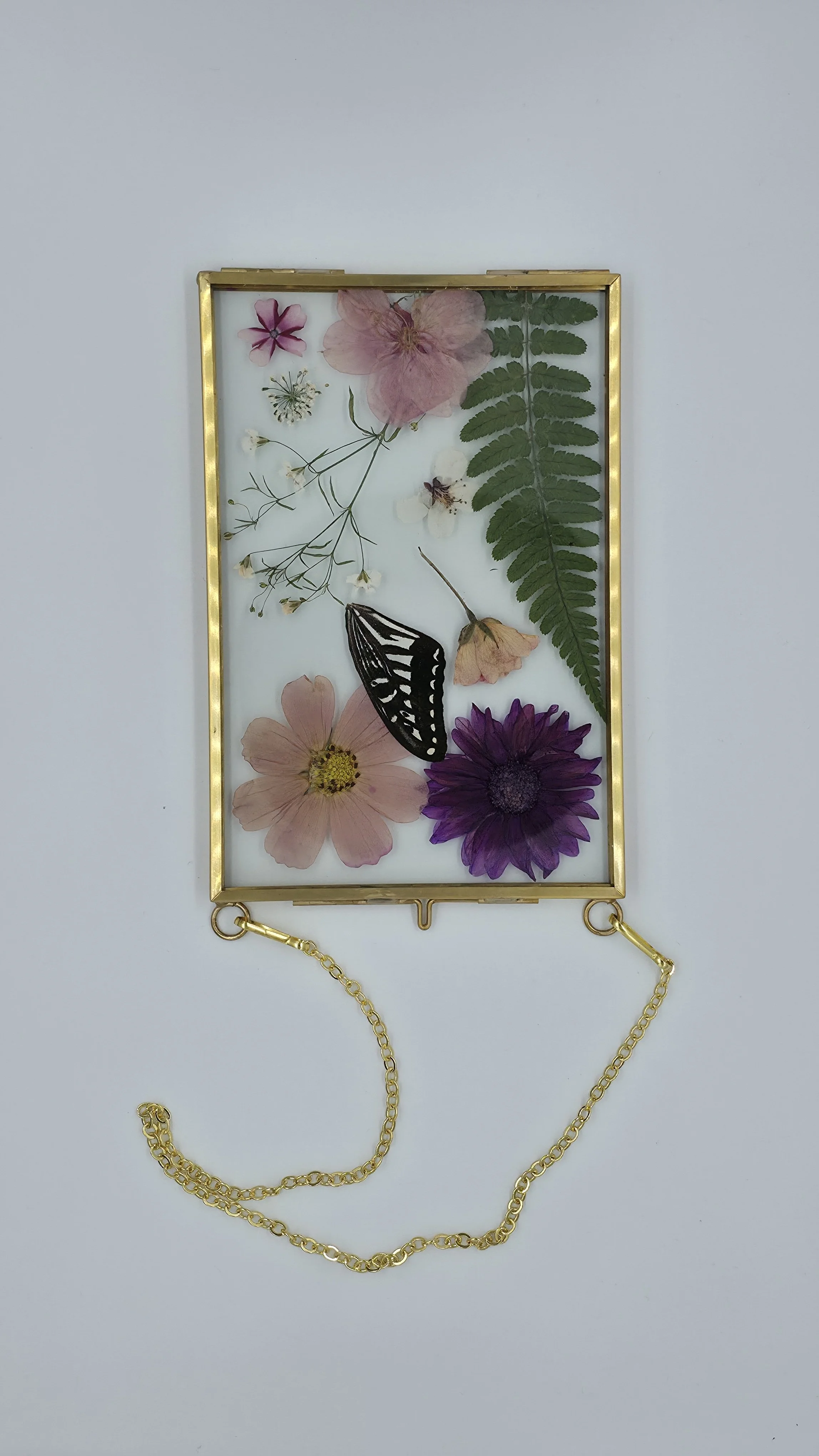 Gold-framed jewelry display with dried pressed flowers, a butterfly, and a gold chain necklace.