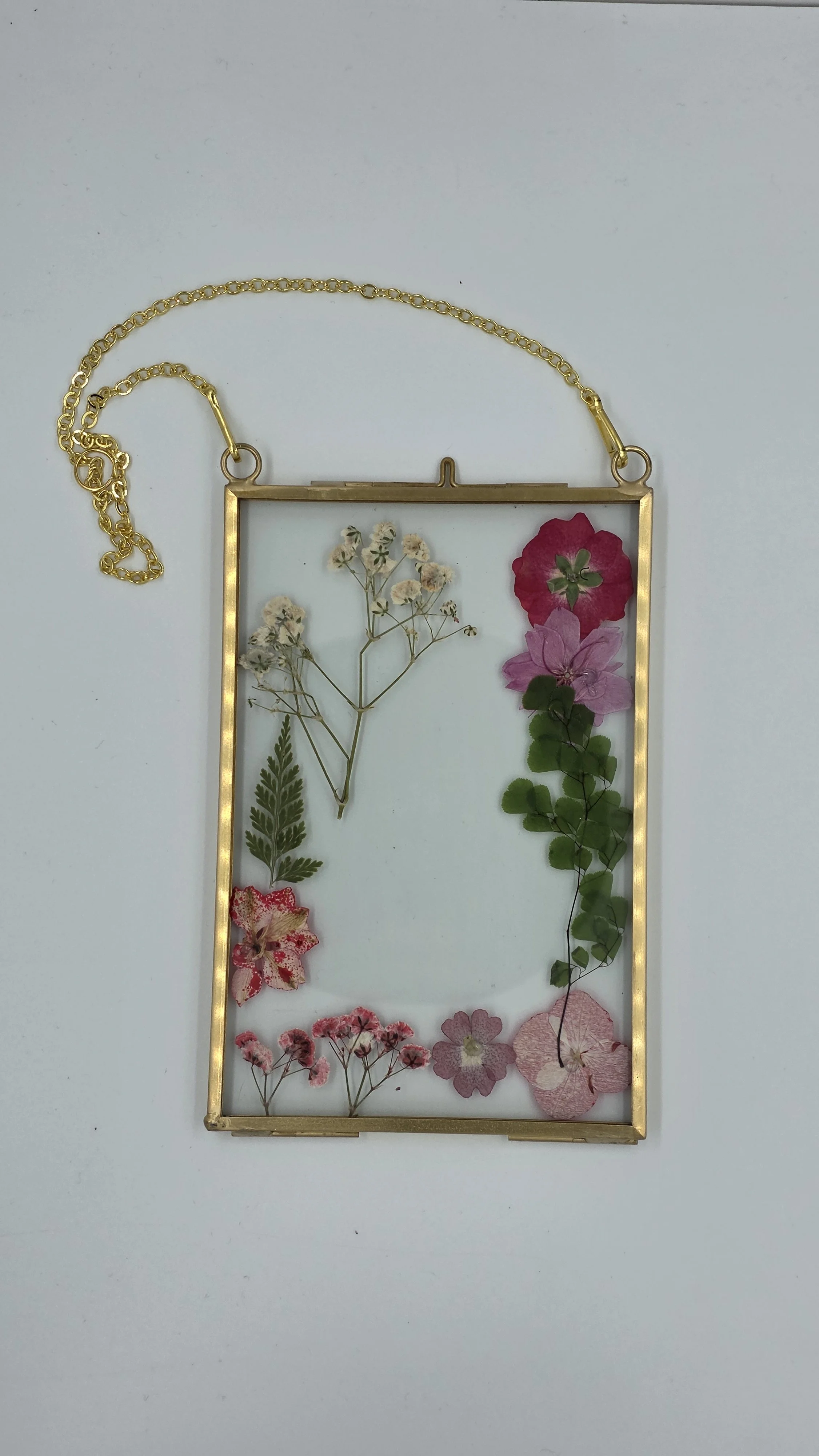 A gold rectangular frame with a chain, containing various pressed dried flowers and leaves arranged artistically.